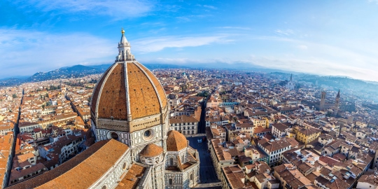 Panoramic aerial view of Florence, Italy, featuring the large red-tiled dome of the Florence Cathedral under a blue sky.