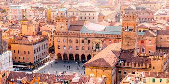 Aerial view of Bologna city center showing historic buildings with terracotta roofs, including the Palazzo d'Accursio with its clock tower and a busy public square beneath.