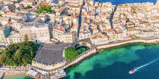 Aerial view of a coastal town with sandy buildings, a stone waterfront promenade, lush green trees, and a small red boat speeding across turquoise water.