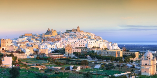 Panoramic view of Ostuni, Italy, showing whitewashed buildings on a hilltop under a soft sunset sky.