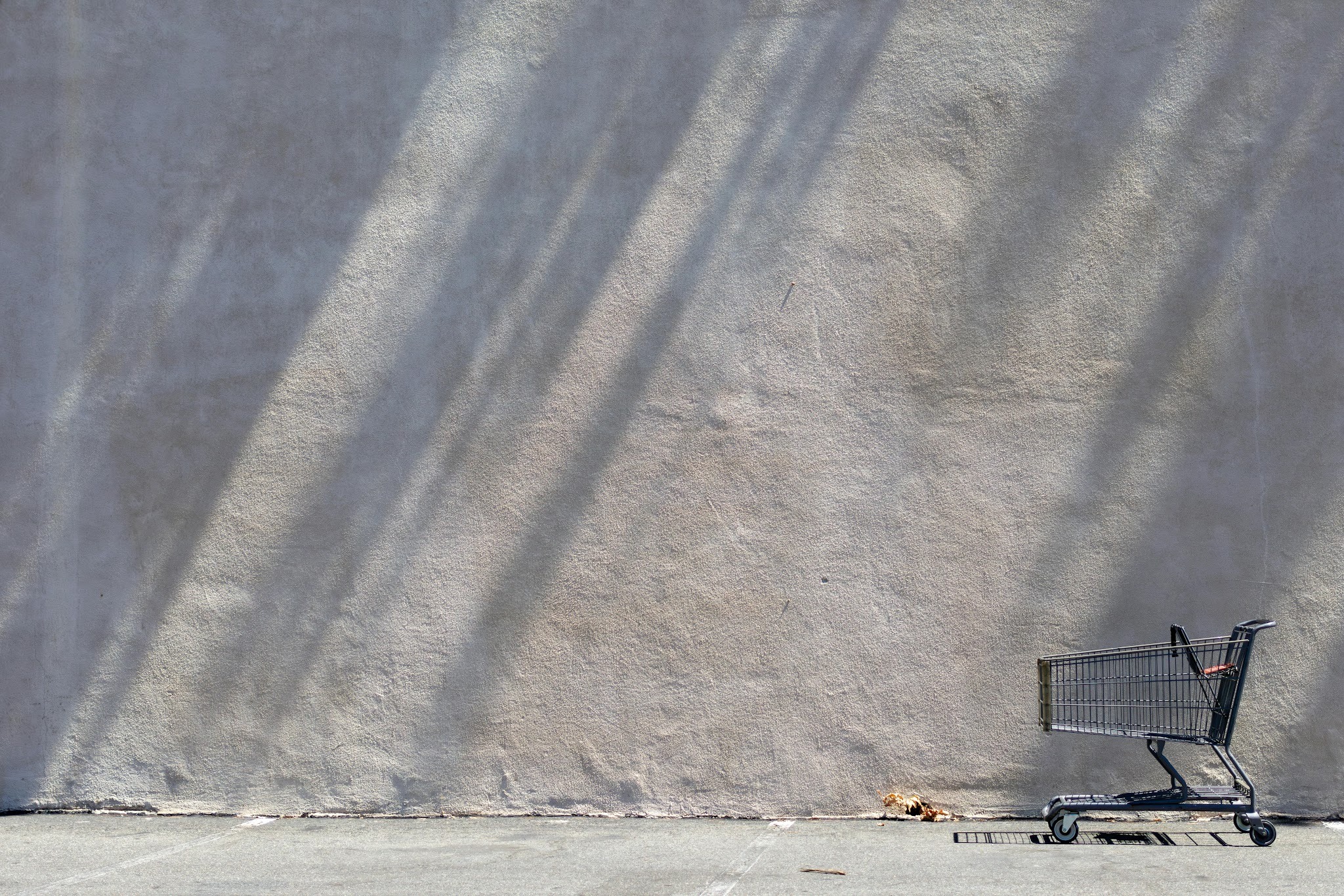 An abandoned shopping cart in a desolate parking lot, pictured against a blank concrete wall.