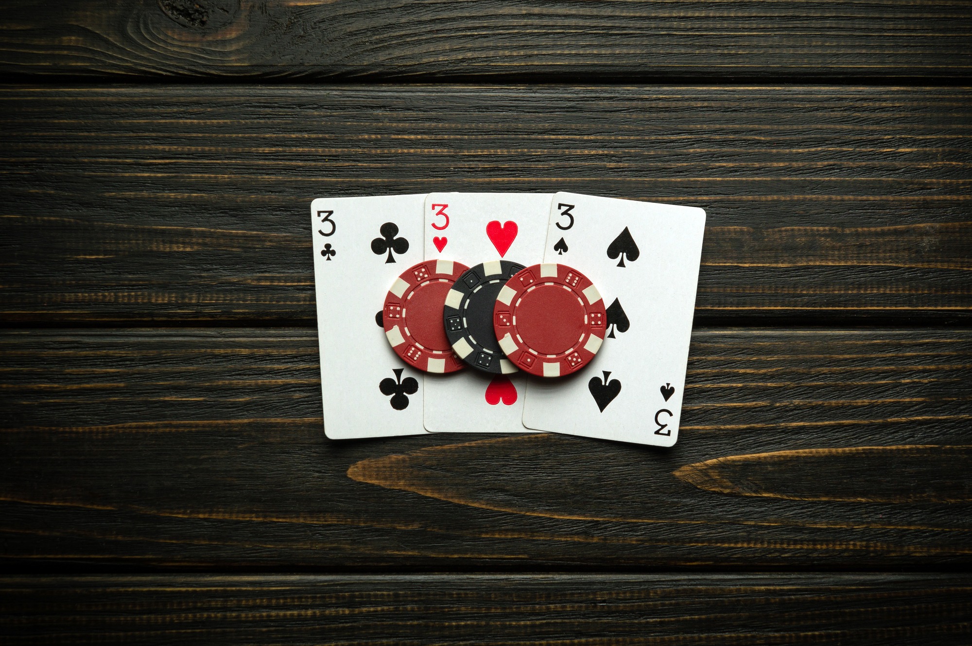 Three playing cards on a table in a matching set, with a three of spades, a three of hearts, and a three of clubs. Three poker chips rest on top of them, one for each card.