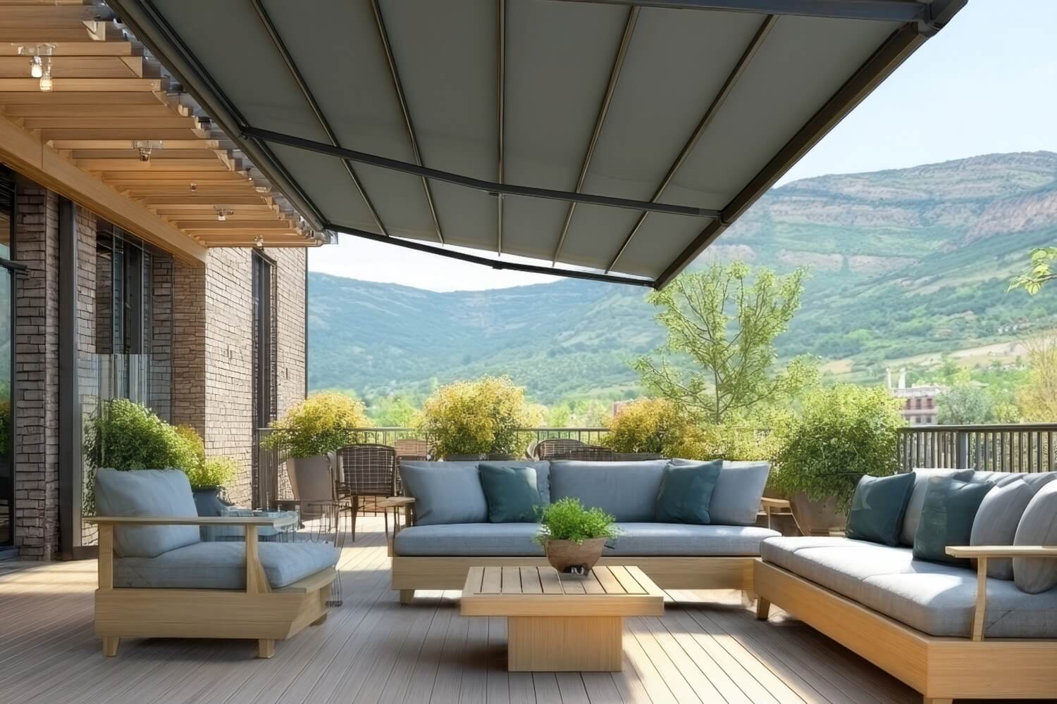 Outdoor terrace with sleek furniture, glass railing, dark awning, built-in sink, and distant view of houses and hills under a partly cloudy sky.