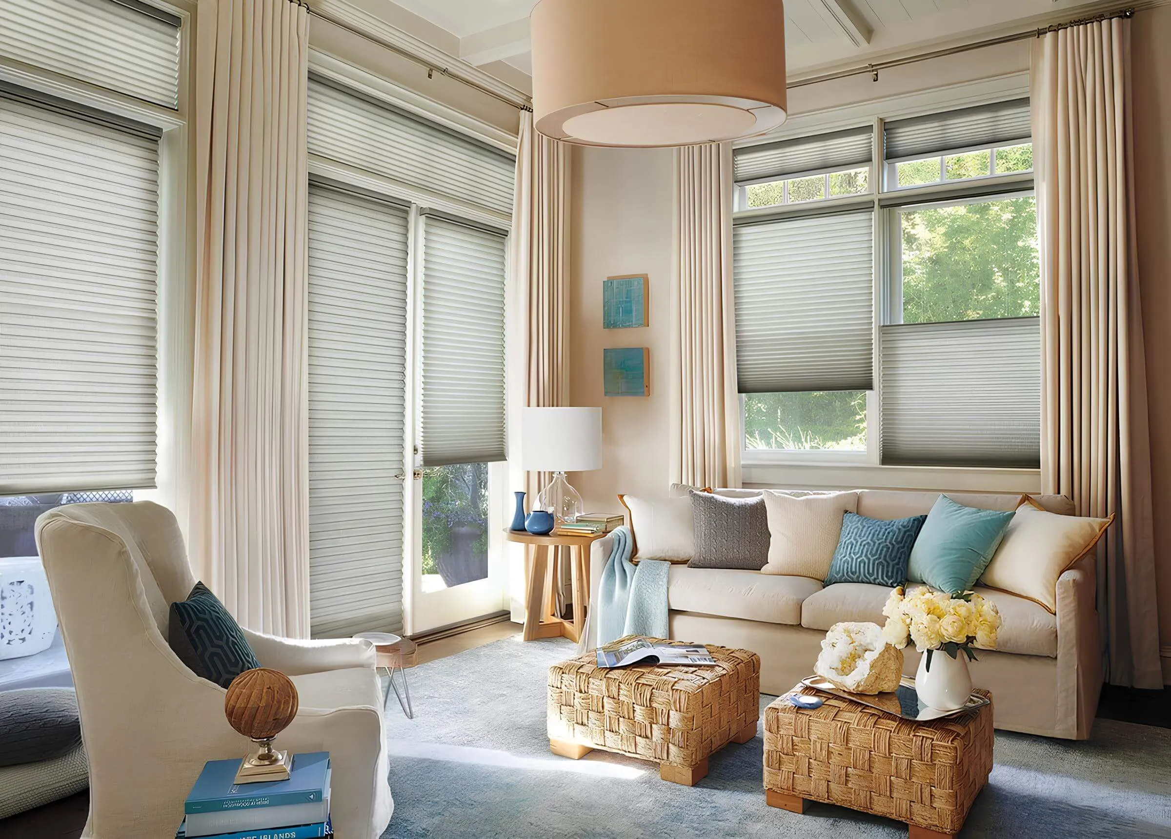 A cozy living room with large windows, blinds, a beige sofa, blue and beige cushions, a wicker coffee table, and a large hanging light fixture.