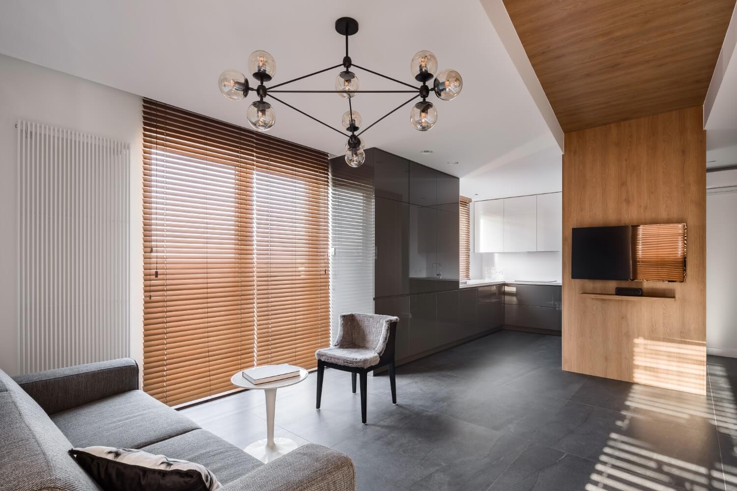 A modern living room with a beige sofa, black armchair, and round wooden coffee table, featuring decorative items and a vase with flowers, set against a background of large windows with horizontal blinds.