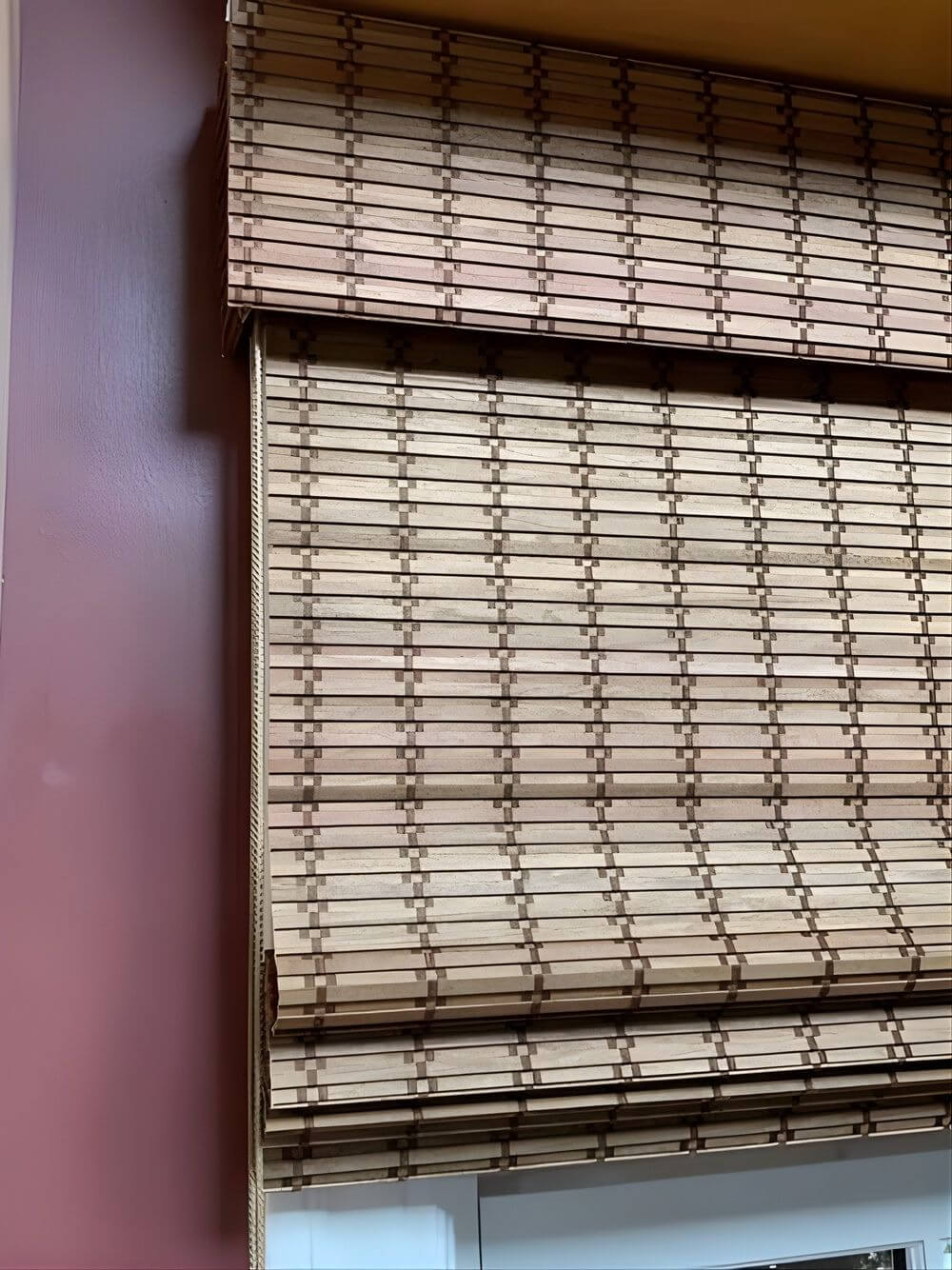 Bamboo window blinds partially rolled up against a maroon wall.
