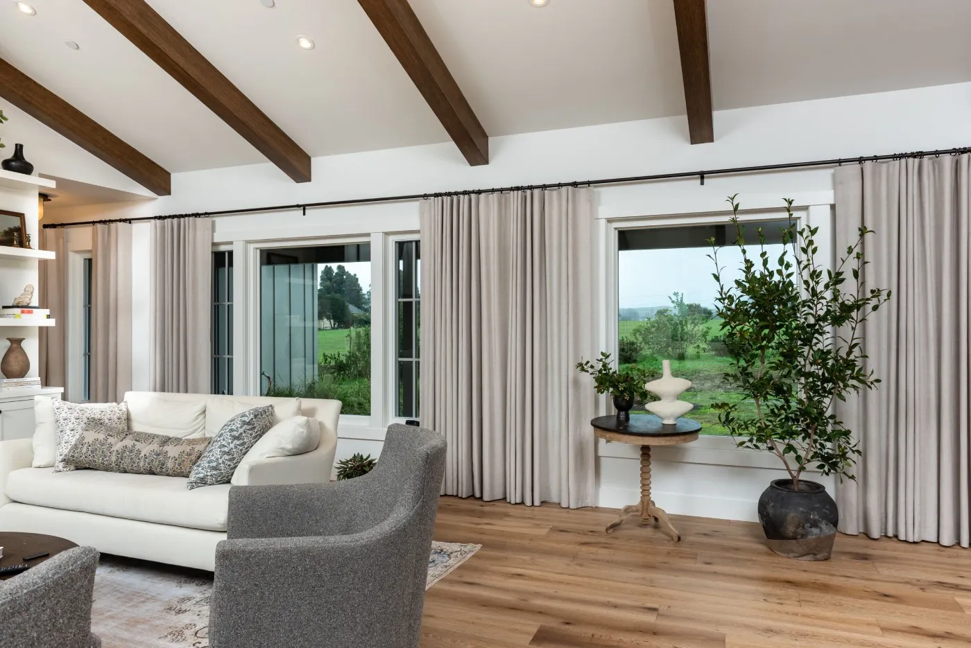 A spacious, modern living room with wooden ceiling beams, a white sofa, large windows with curtains, and a potted plant on the side.