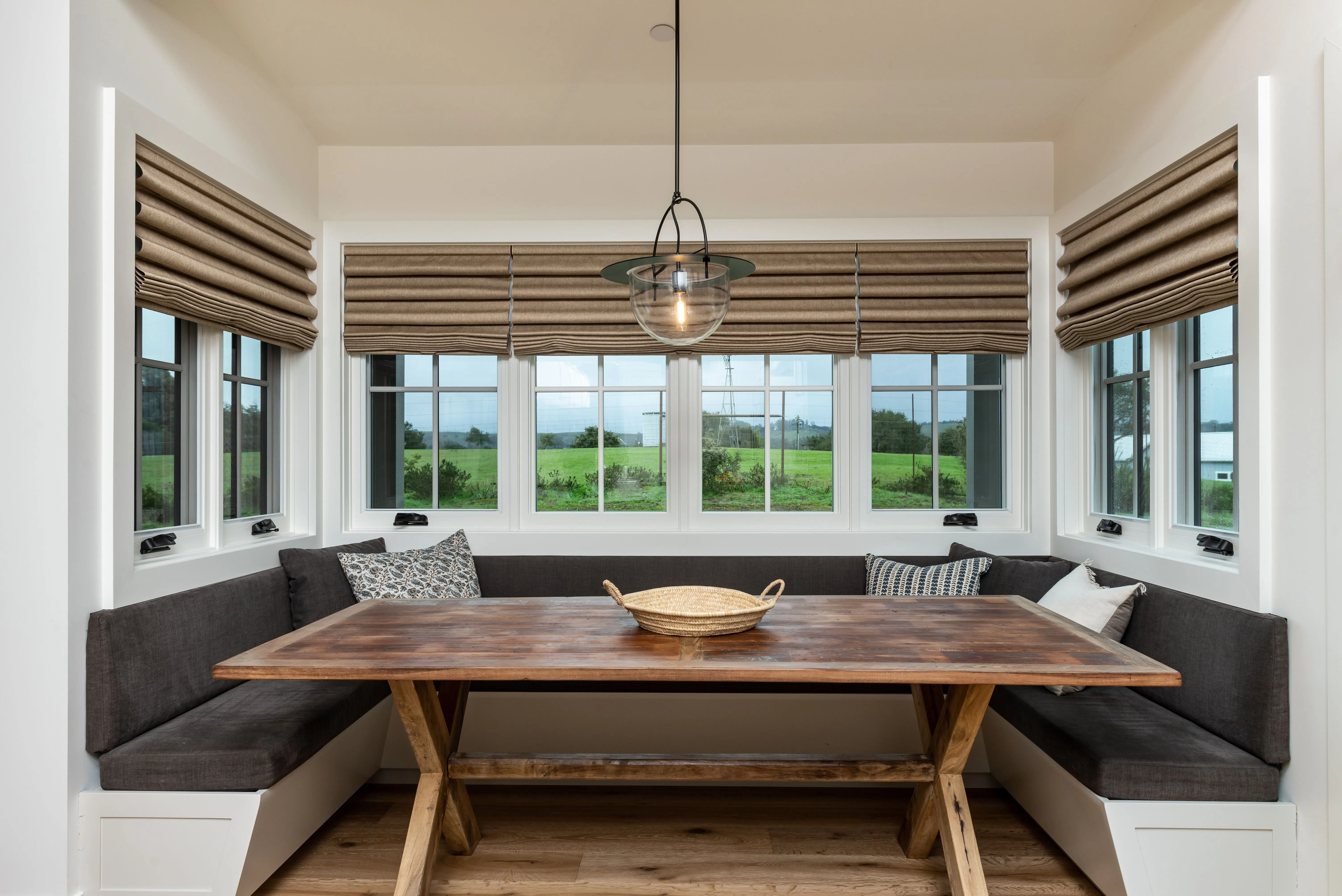 Cozy dining nook with a wooden table, gray cushioned benches, striped window shades, and a hanging light fixture, overlooking a green outdoor view.