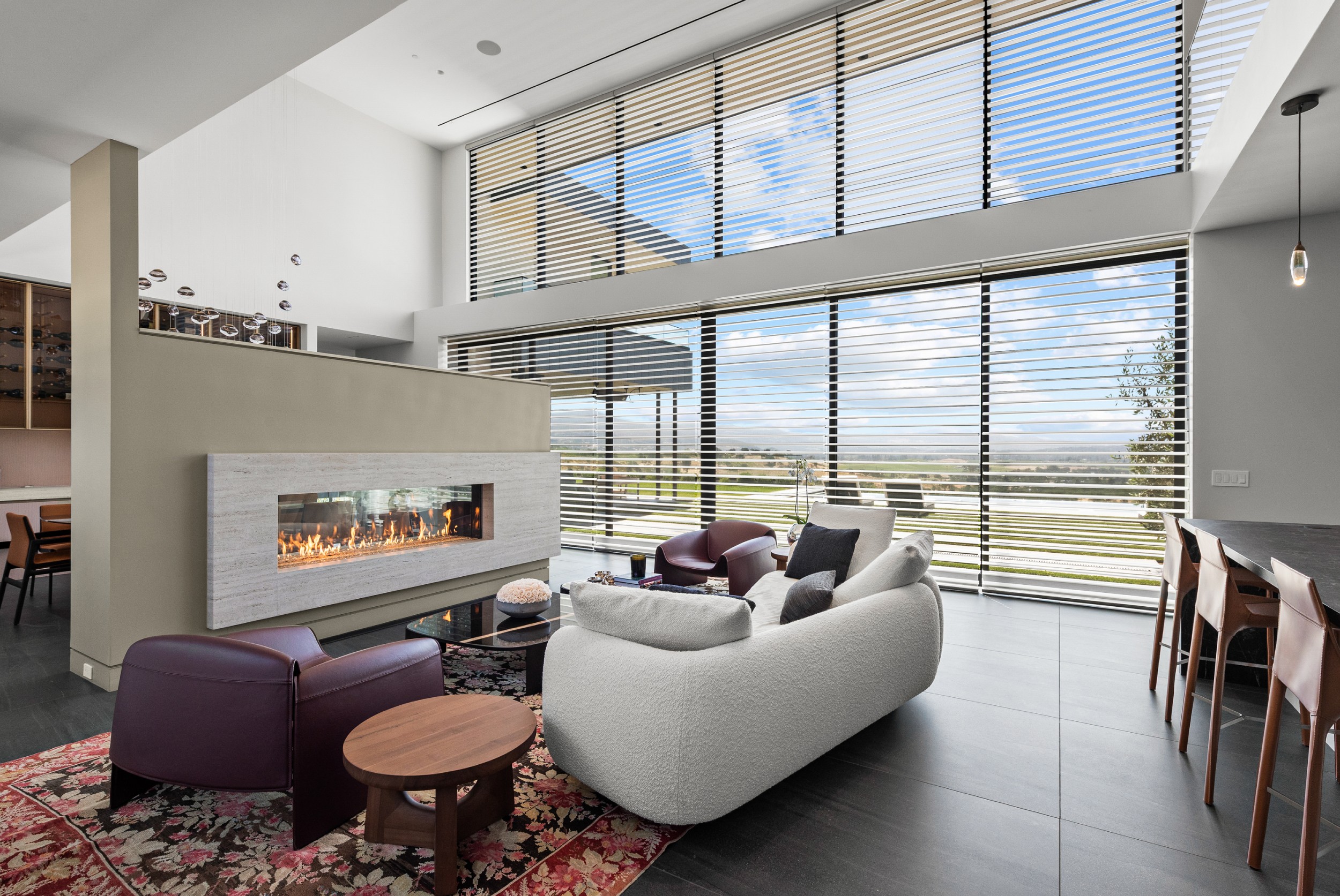Modern living room with floor-to-ceiling windows covered by horizontal wooden blinds, allowing natural light to filter through while offering a view of the landscape outside.