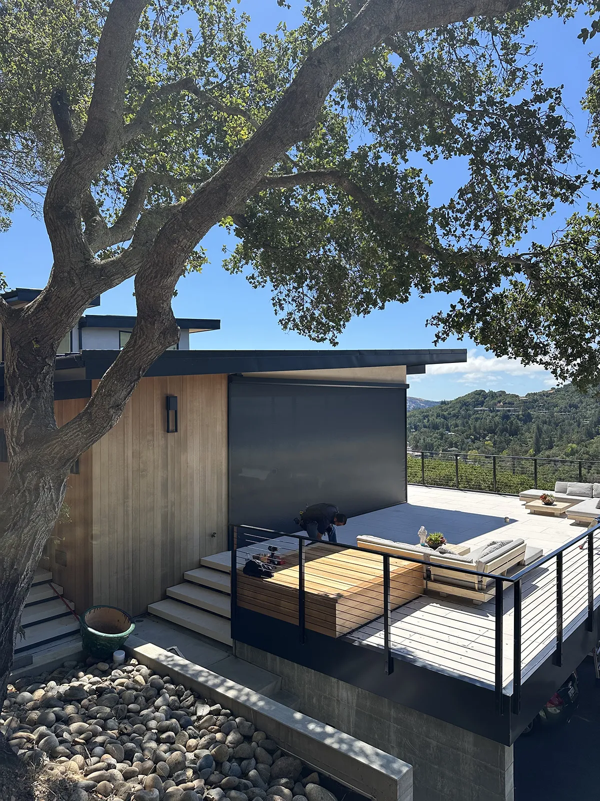 Modern wooden deck with seating, surrounded by trees, overlooking a scenic mountain landscape under a clear sky.