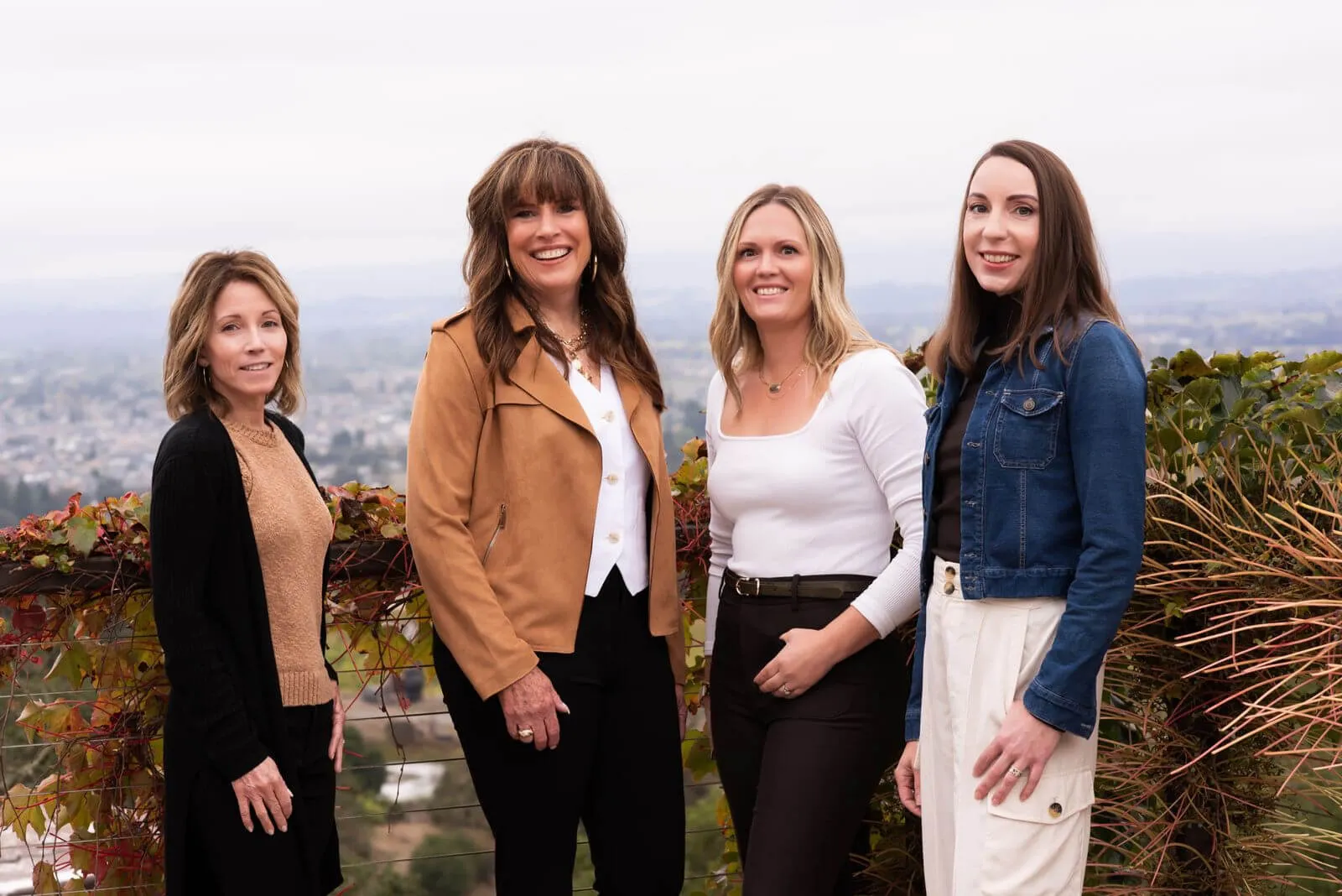 Four smiling women standing outdoors in front of a leafy railing with a cityscape in the background.