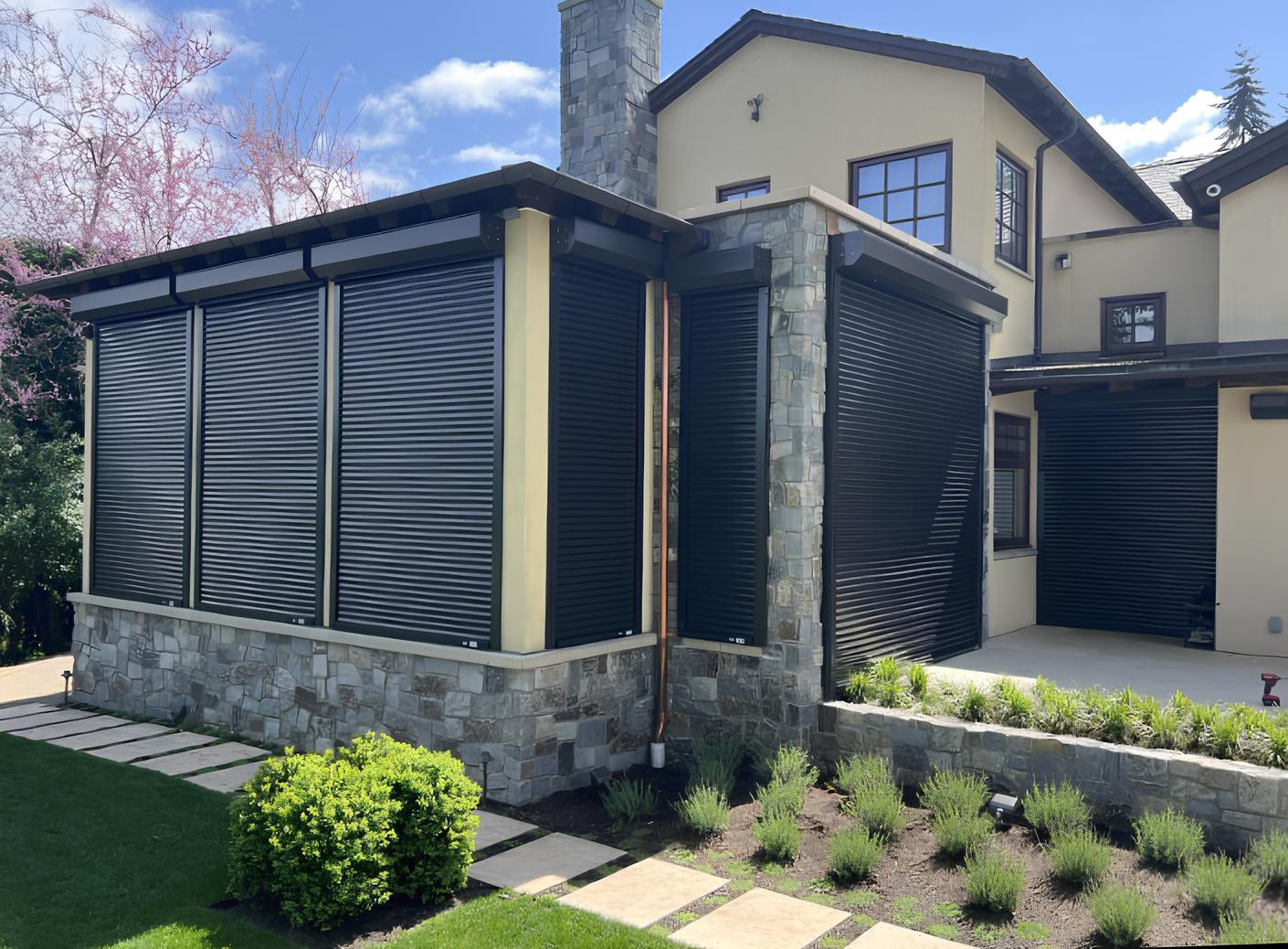House with stone base and black security roll-up shutters on its windows and doors on a sunny day.