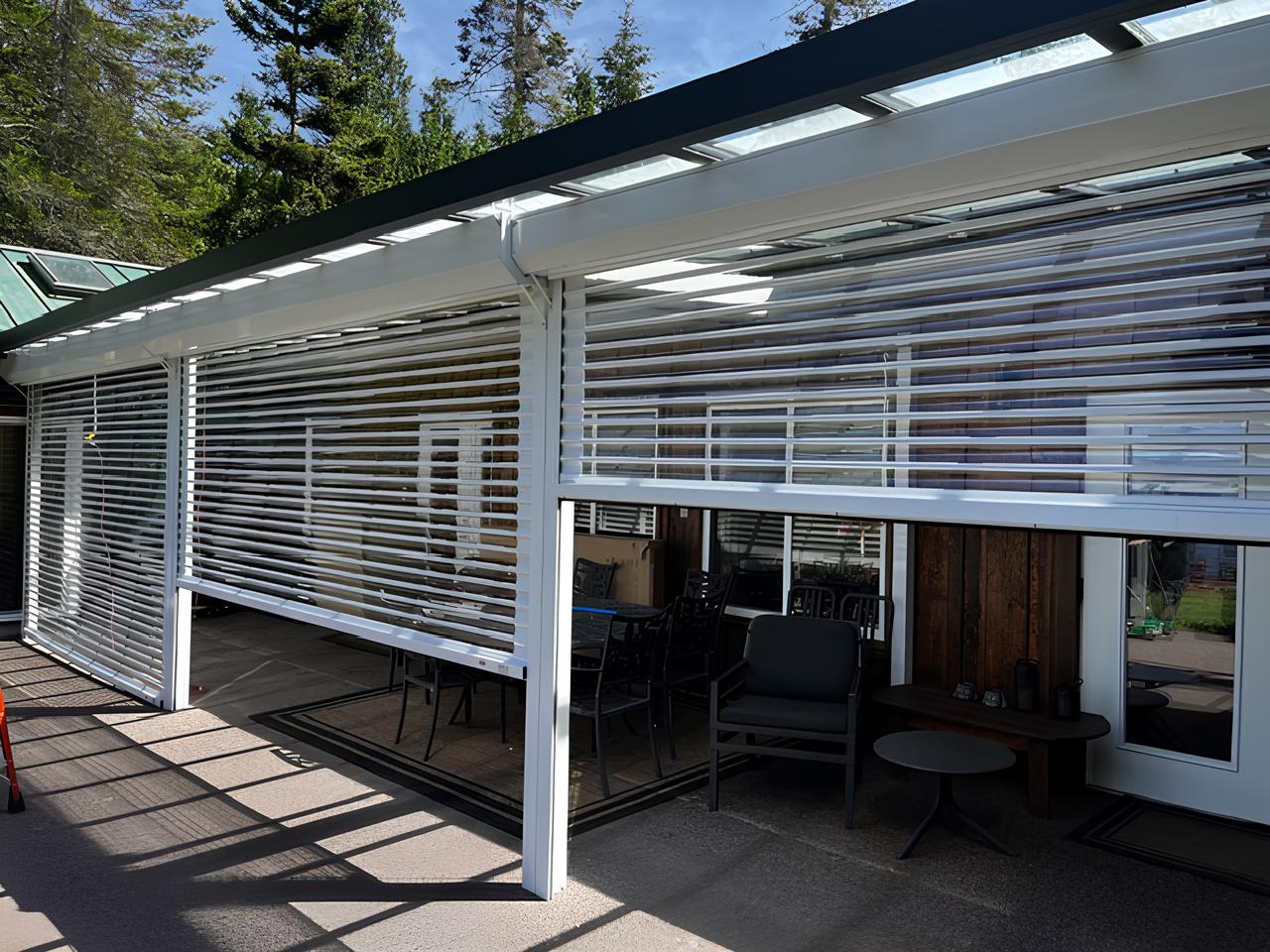 Outdoor patio with seating behind partially lowered white horizontal slat blinds under a transparent roof.