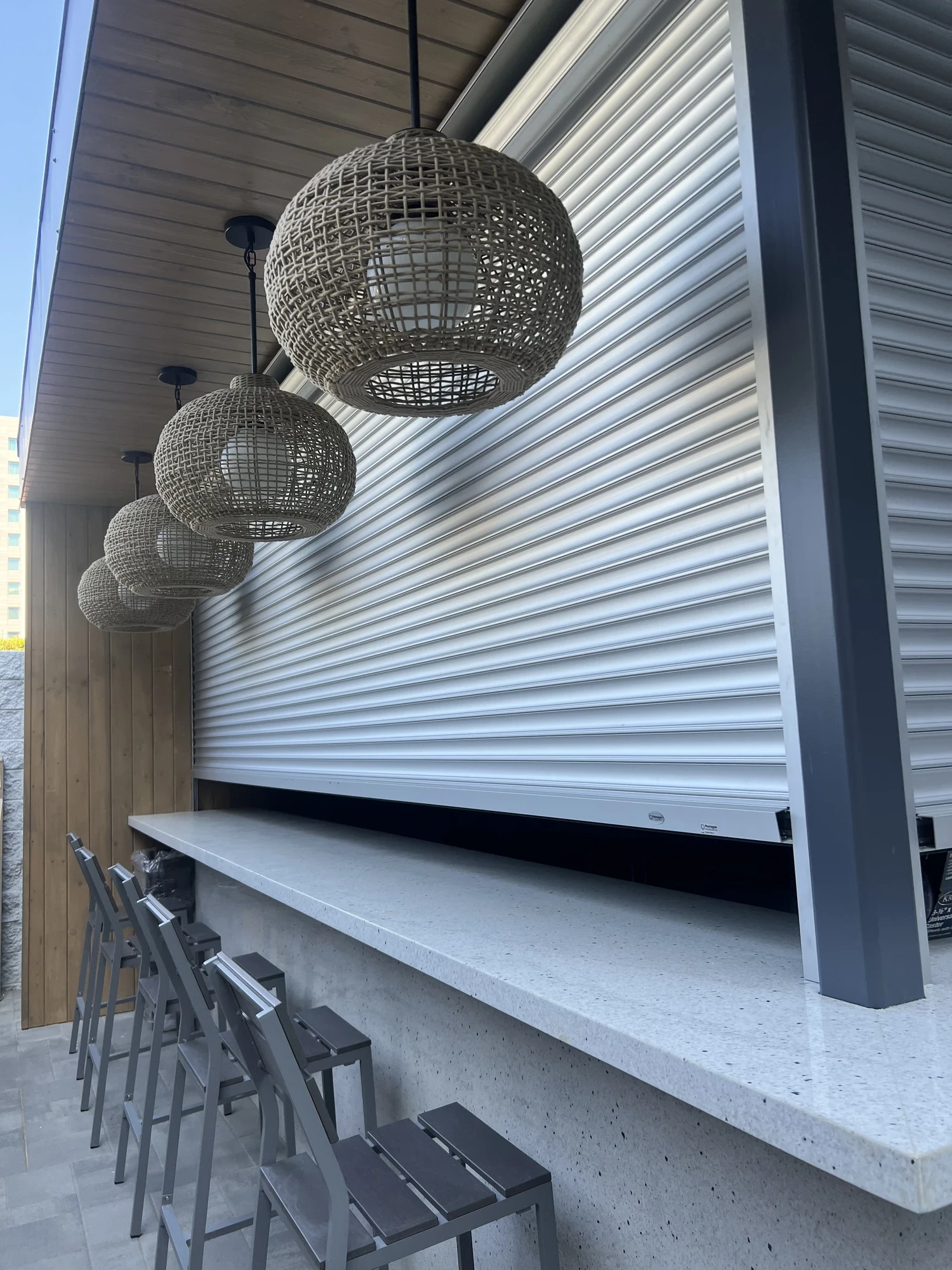 Outdoor bar with gray countertop, metal stools, hanging wicker pendant lights, and a closed metal shutter.
