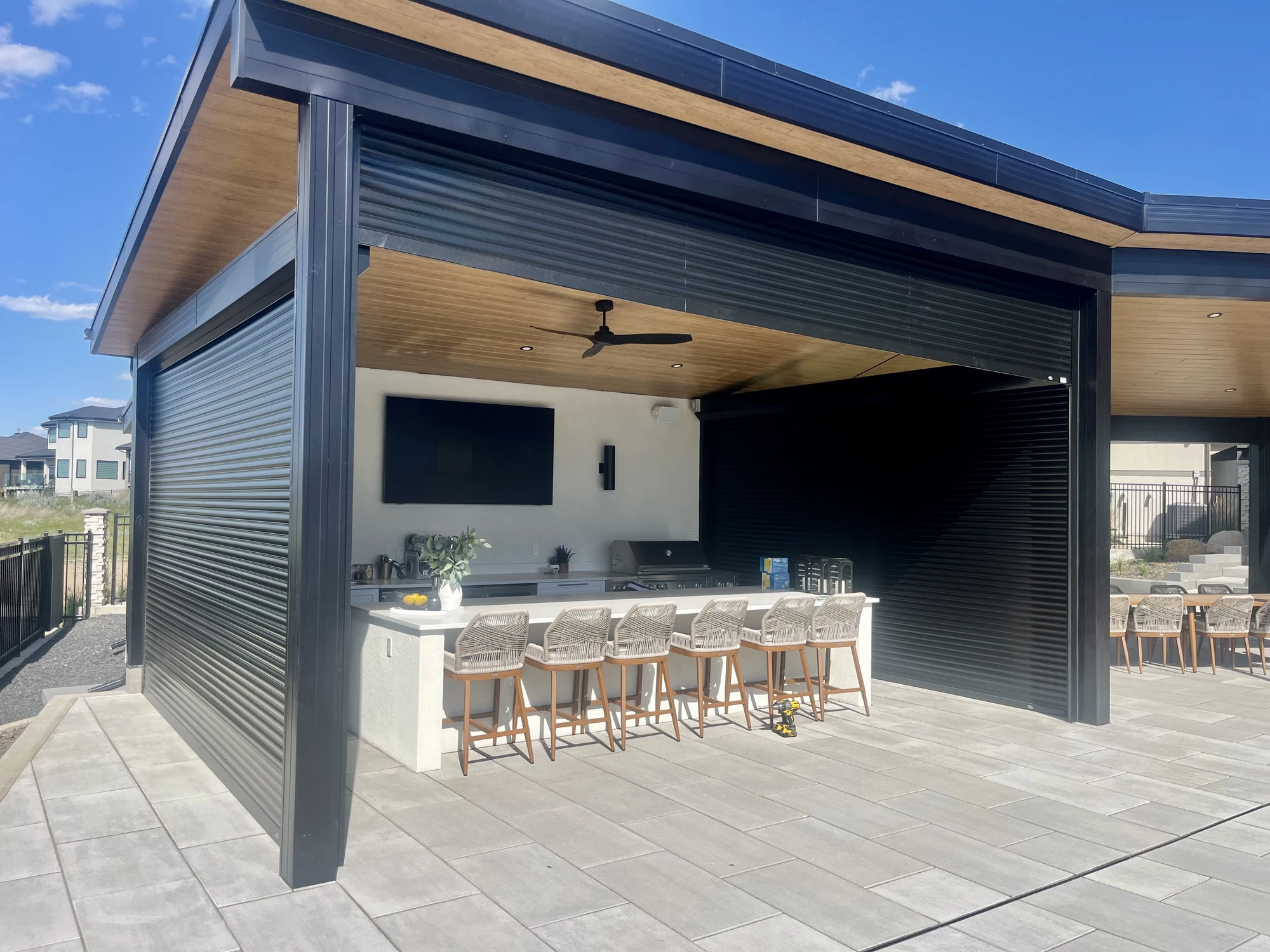 Modern outdoor kitchen with bar stools, ceiling fan, TV, and metal roll-up garage doors.