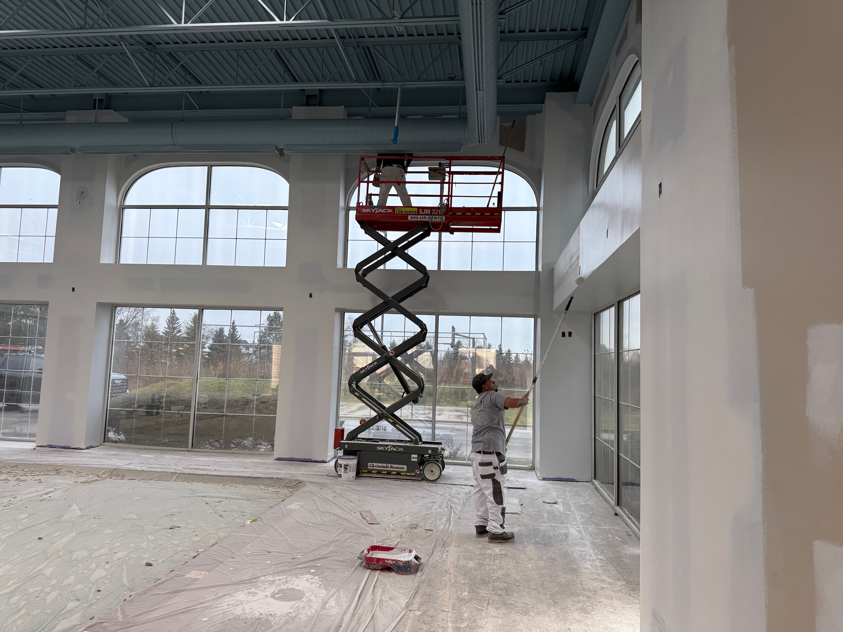 Painter rolling fresh white paint onto upper pool room wall under large windows.