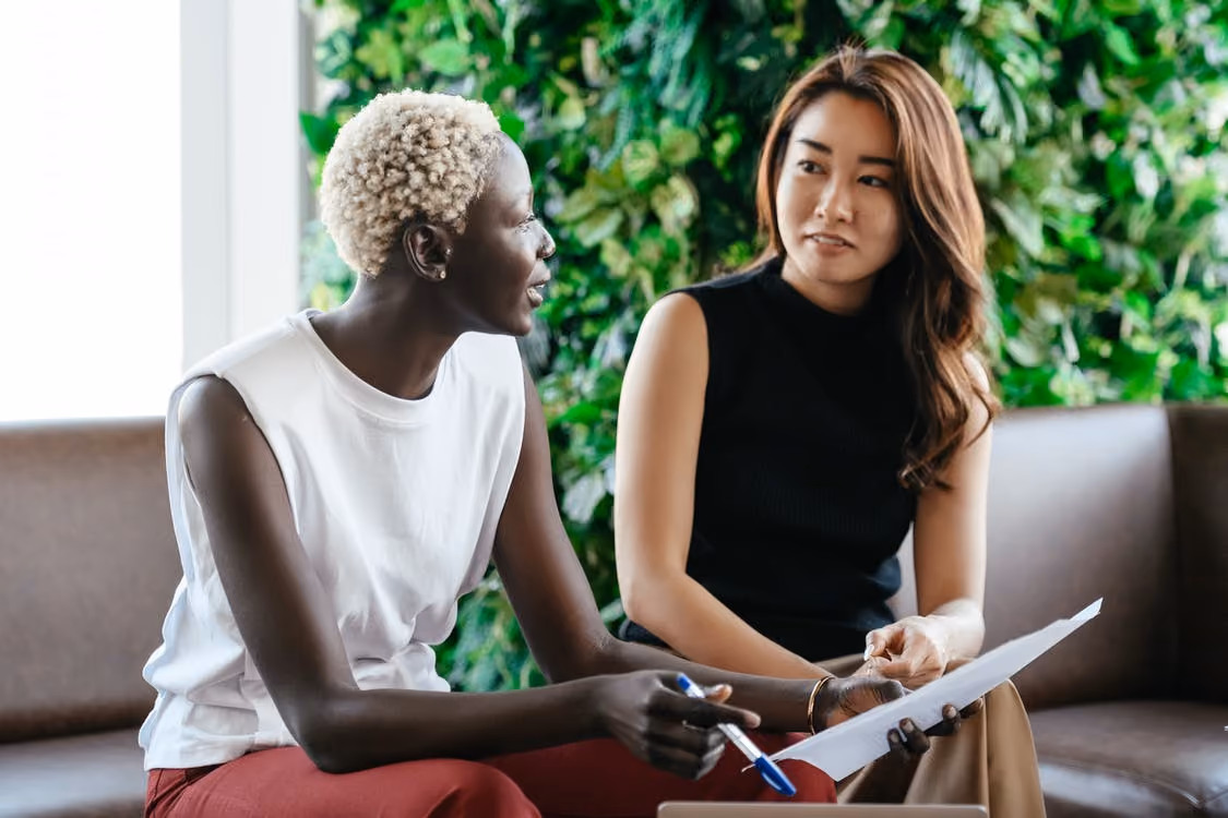 Two women sitting on a brown couch having a discussion; one woman with short curly blonde hair wearing a white sleeveless top and red pants holds a pen and paper, while the other woman with long brown hair wearing a black sleeveless top listens attentively against a backdrop of green leafy plants.