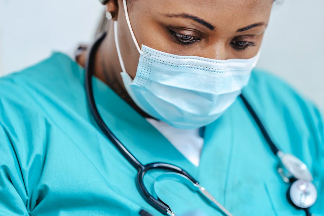 Healthcare professional wearing teal scrubs, a face mask, and a stethoscope around the neck, looking down attentively.