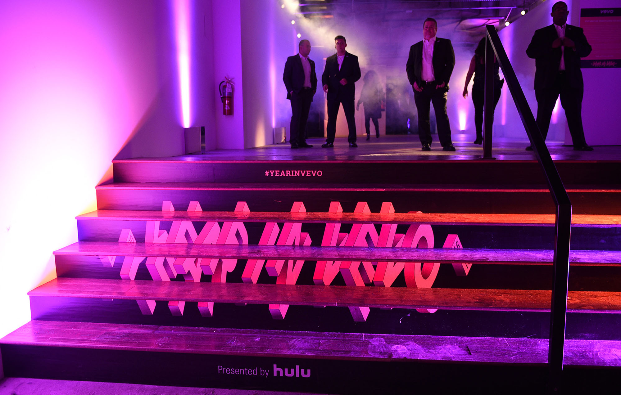 a stair case with the vevo in a year logo on them and security guards wearing suits on top of the stair case with purple light in the background
