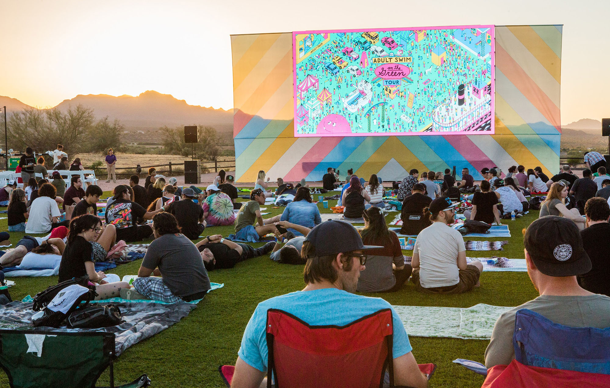 people on a green lawn sitting on beach towel in front of a large screen 