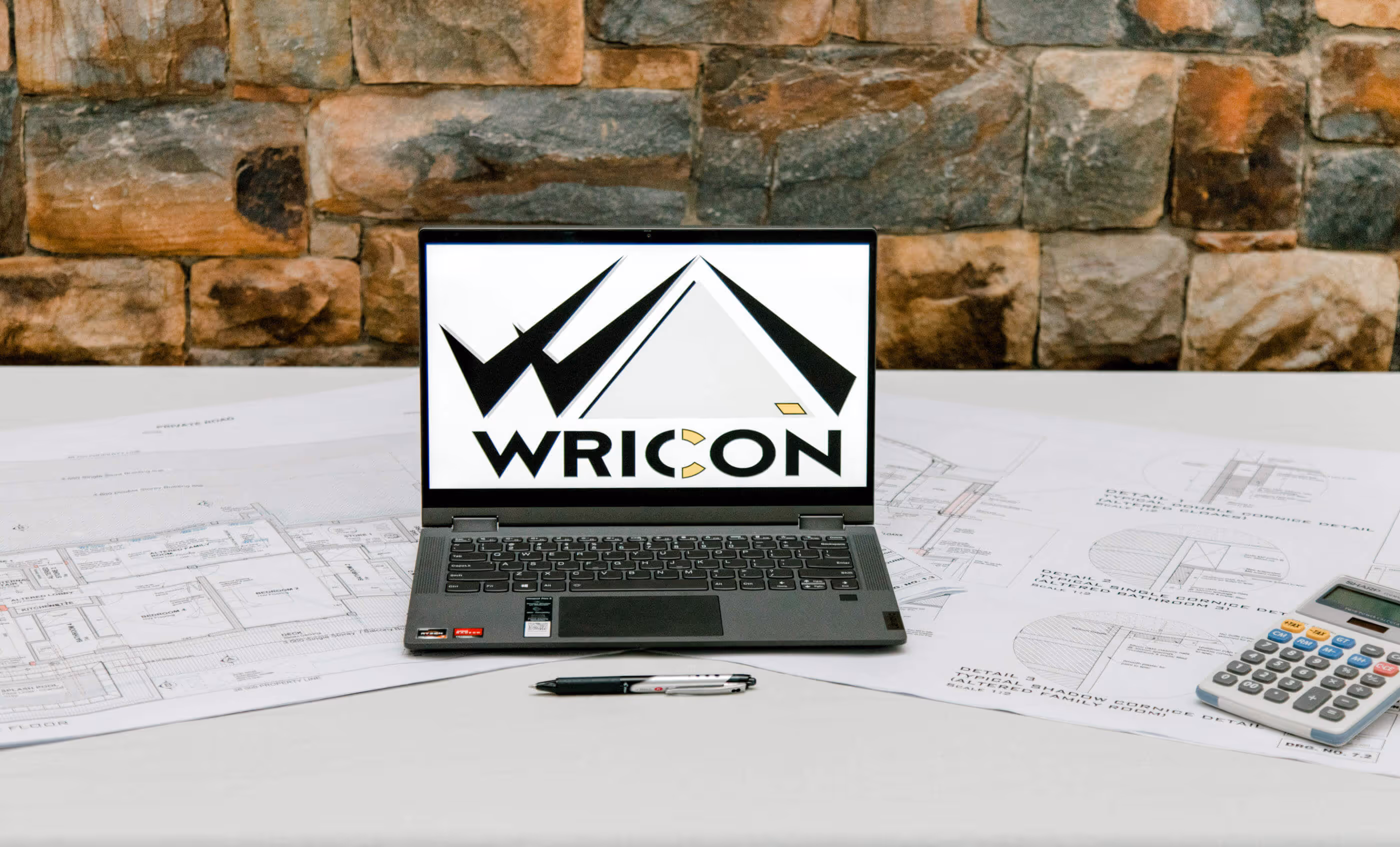 Laptop displaying a WRICON logo on the screen, placed on architectural blueprints with a calculator and pen nearby, against a stone wall background.