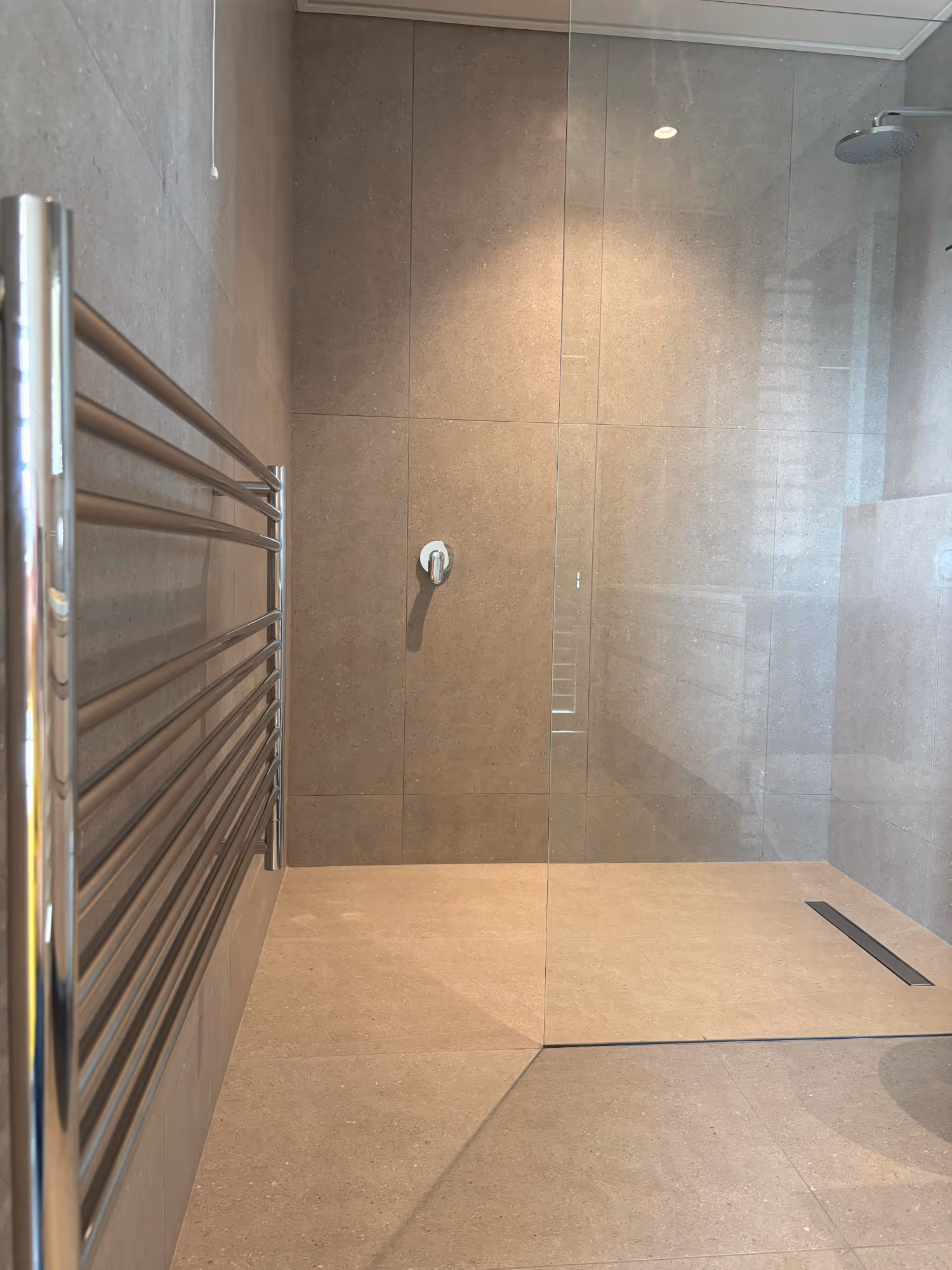 Modern walk-in shower with beige tile walls and floor, glass partition, overhead rain shower, and chrome towel radiator. Work by Wricon construction.