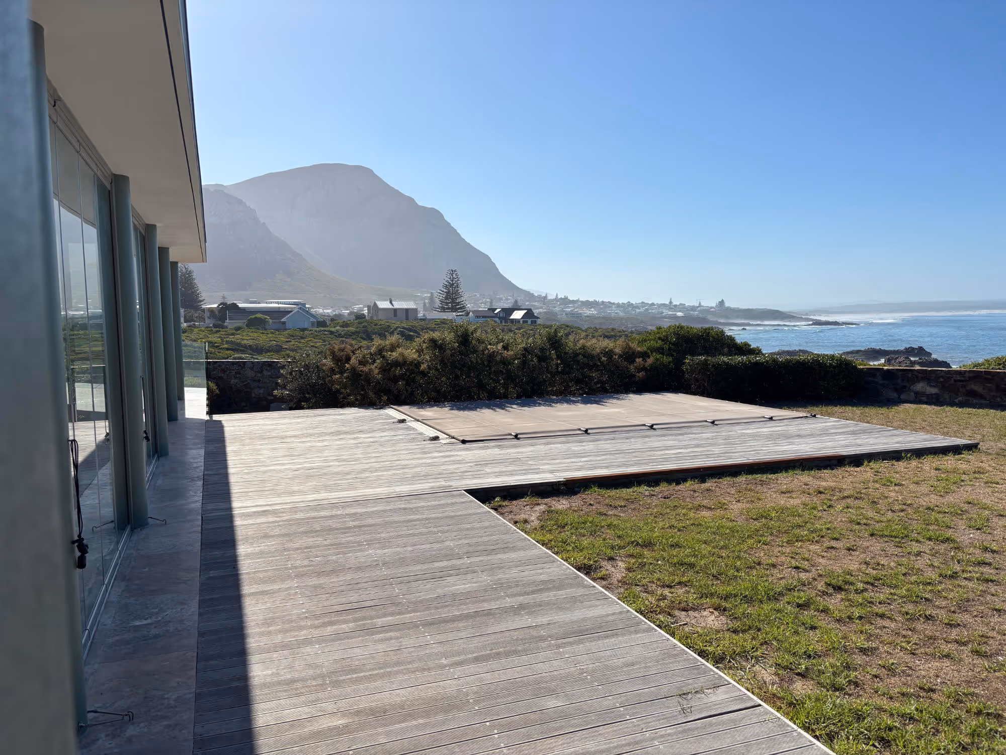 Modern wooden deck next to a house with glass walls overlooking greenery, distant mountains, and the ocean under a clear blue sky. Work by Wricon construction.