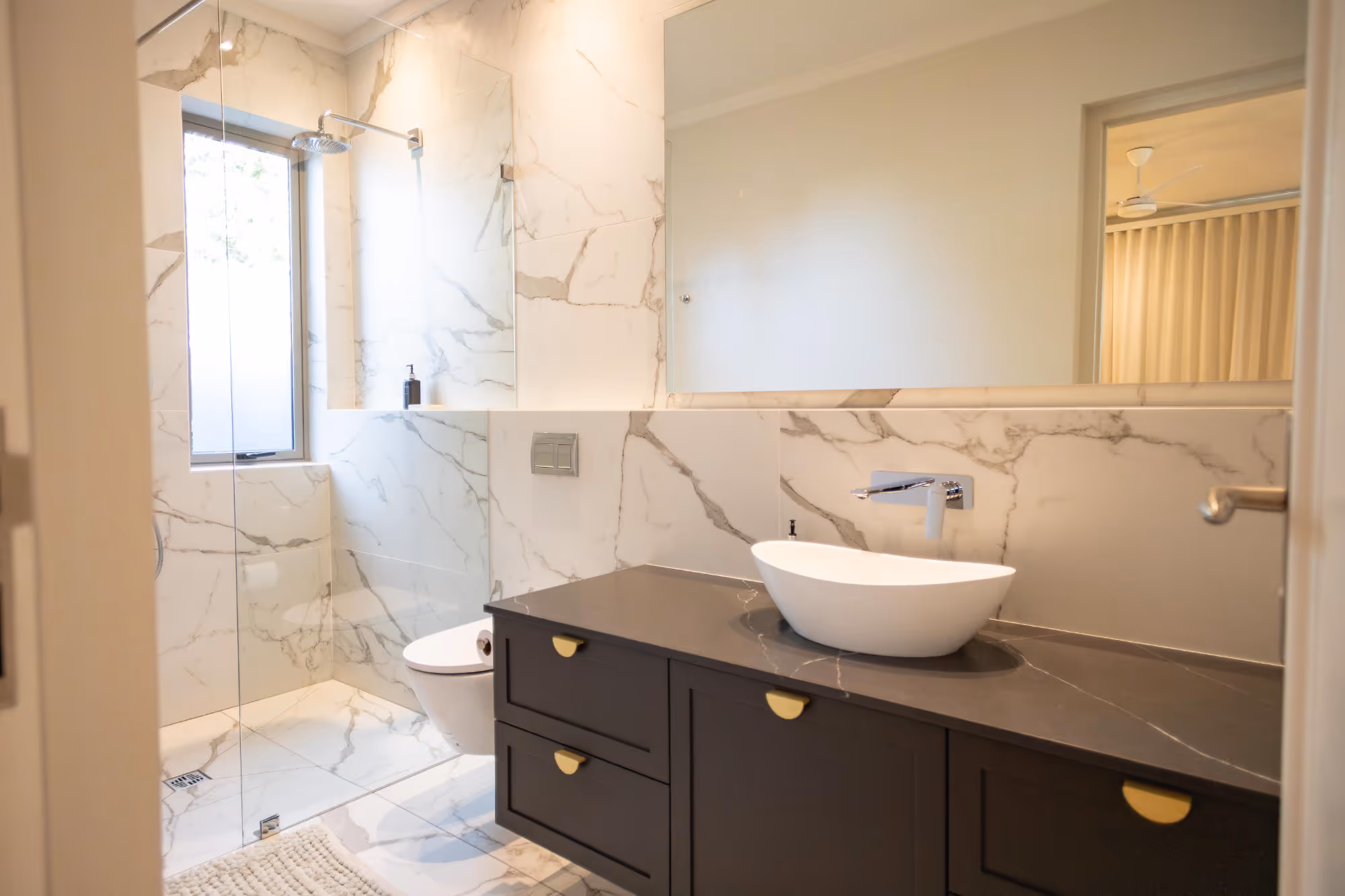 Modern bathroom with marble walls, a glass-enclosed shower, floating toilet, dark vanity with white vessel sink, and large mirror. Work by Wricon construction.