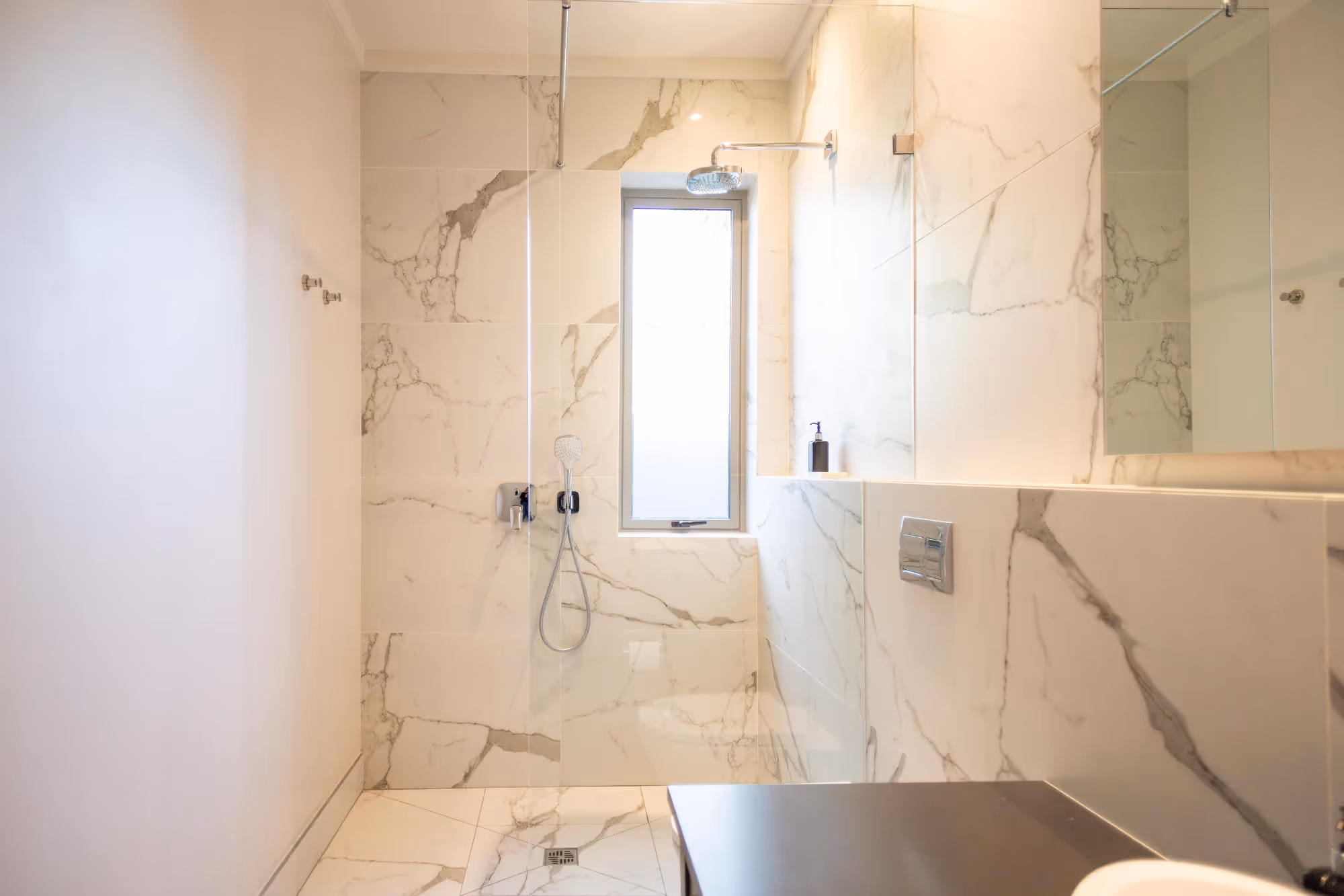 Modern bathroom shower area with large marble tiles, glass partition, rain shower head, handheld shower, and a narrow frosted window. Work by Wricon construction.