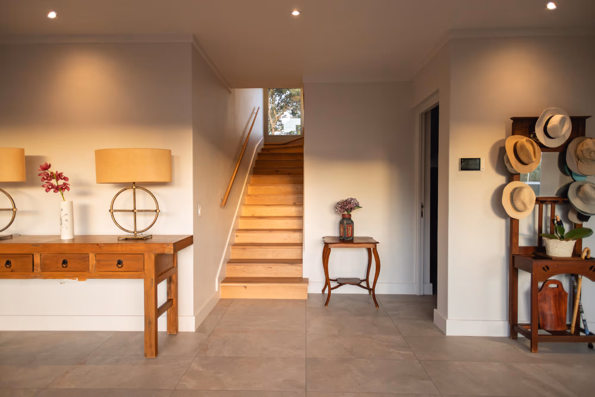 Bright entryway with wooden stairs, a wooden console table with lamps and flowers, and a hat rack with multiple hats. Work by Wricon construction.