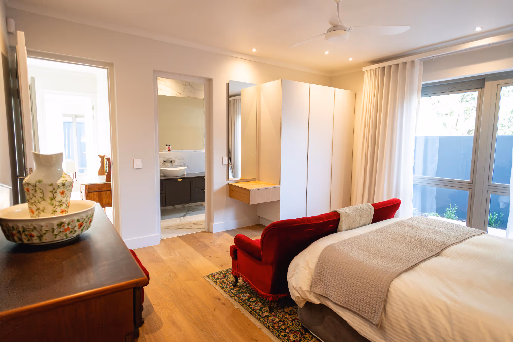 Bright bedroom with a large bed covered in white and gray bedding, a red velvet loveseat at the foot of the bed, wooden floor, white wardrobe, and an open doorway leading to a bathroom. Work by Wricon construction.
