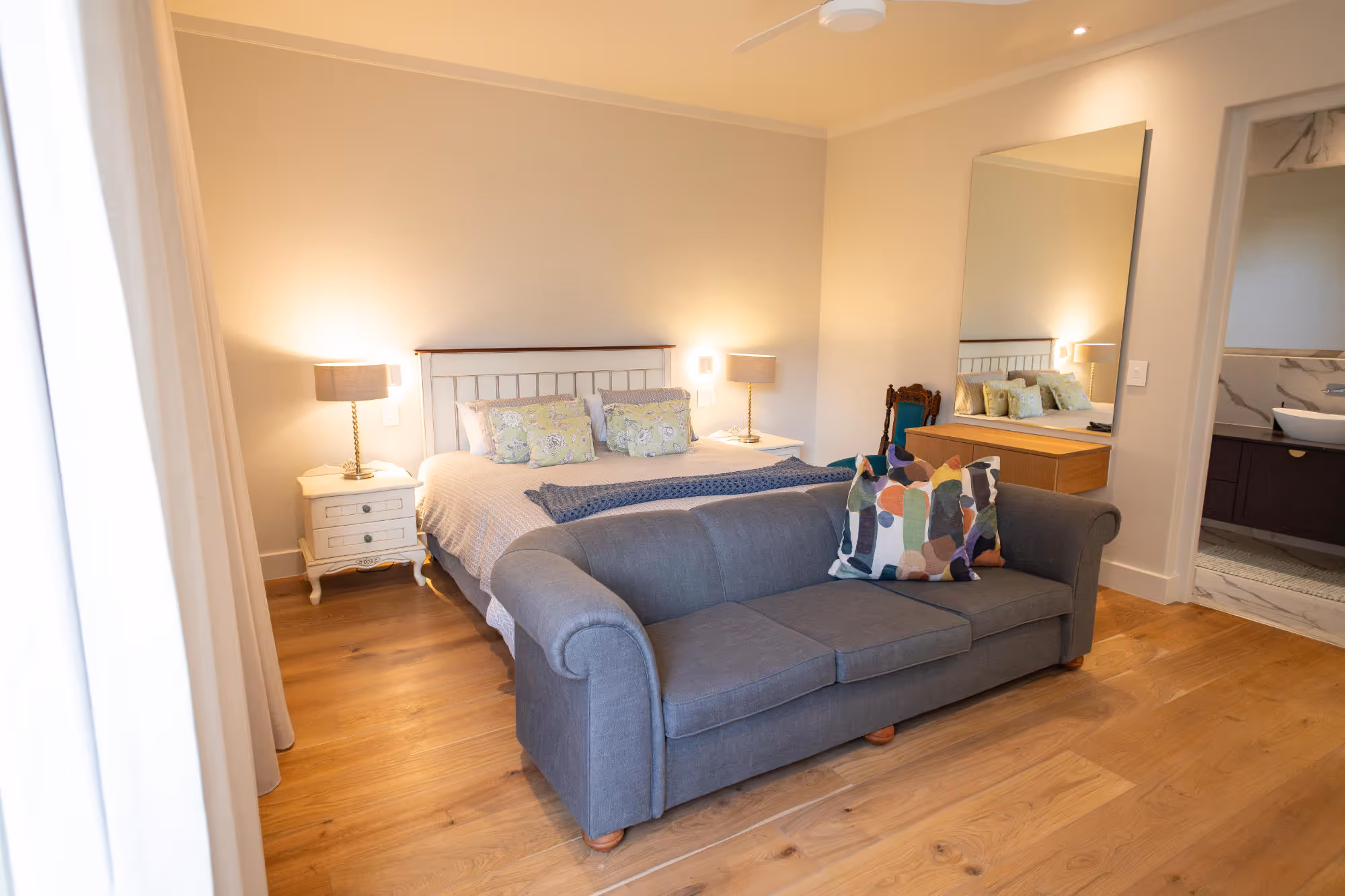 Cozy bedroom with a double bed, two bedside tables with lamps, a gray sofa with colourful pillows, wooden floor, and a large mirror reflecting the bed. Work by Wricon construction.