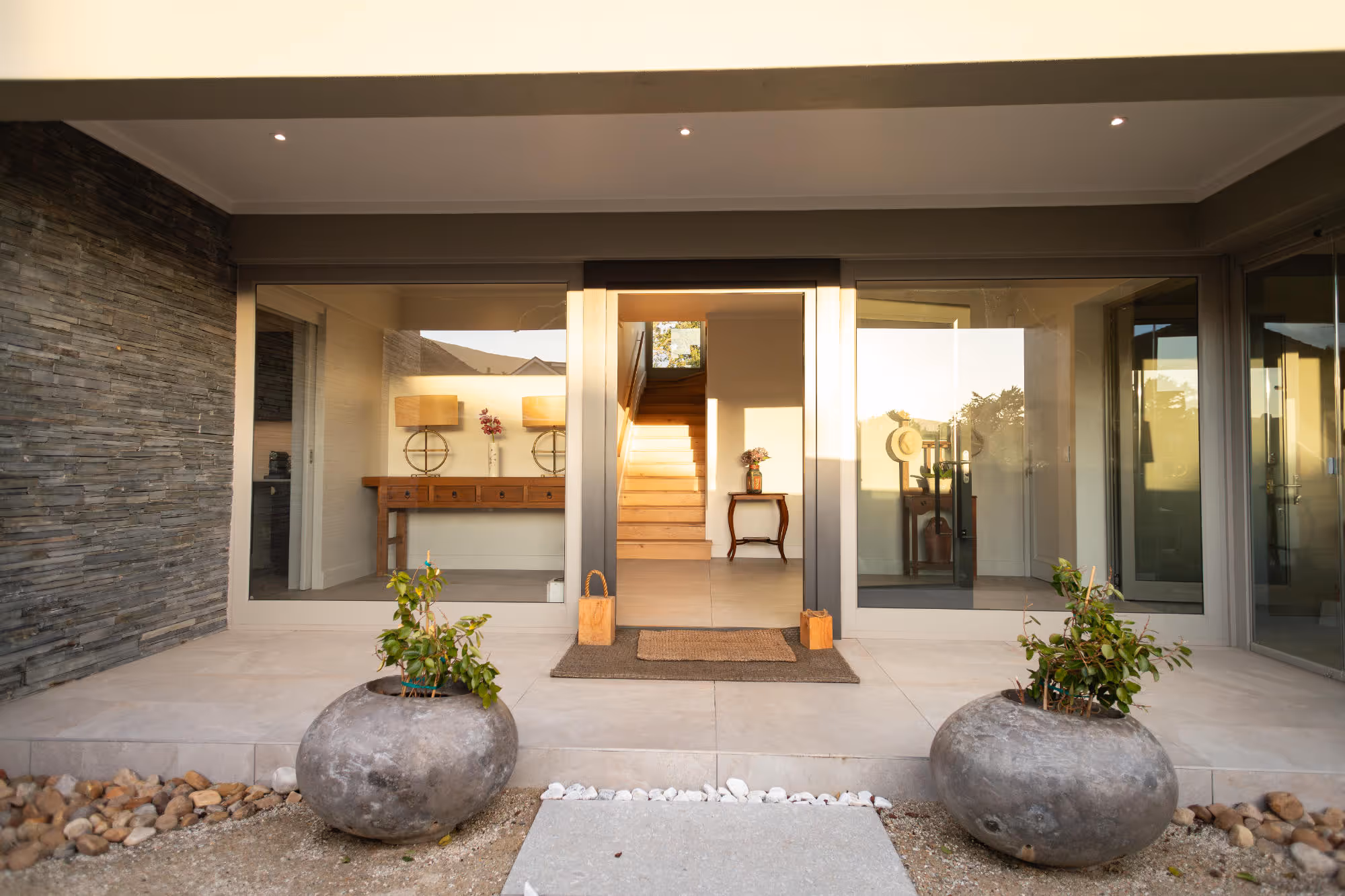 Modern house entrance with large glass doors, two round gray planters, and wooden stairs inside. Work by Wricon construction.