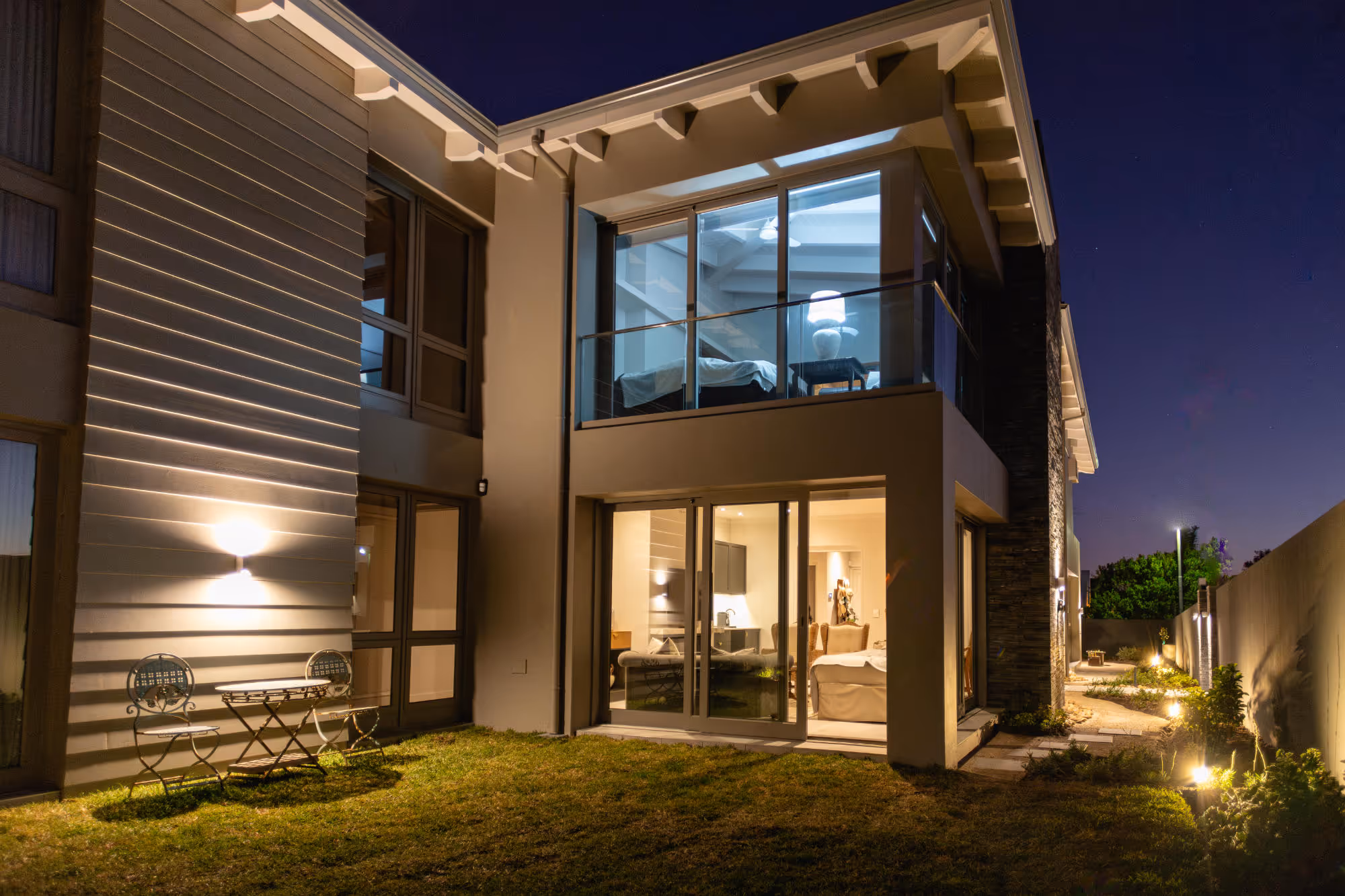 Modern two-story house at night with illuminated interior, glass balcony, and outdoor patio furniture on grass. Work by Wricon construction.