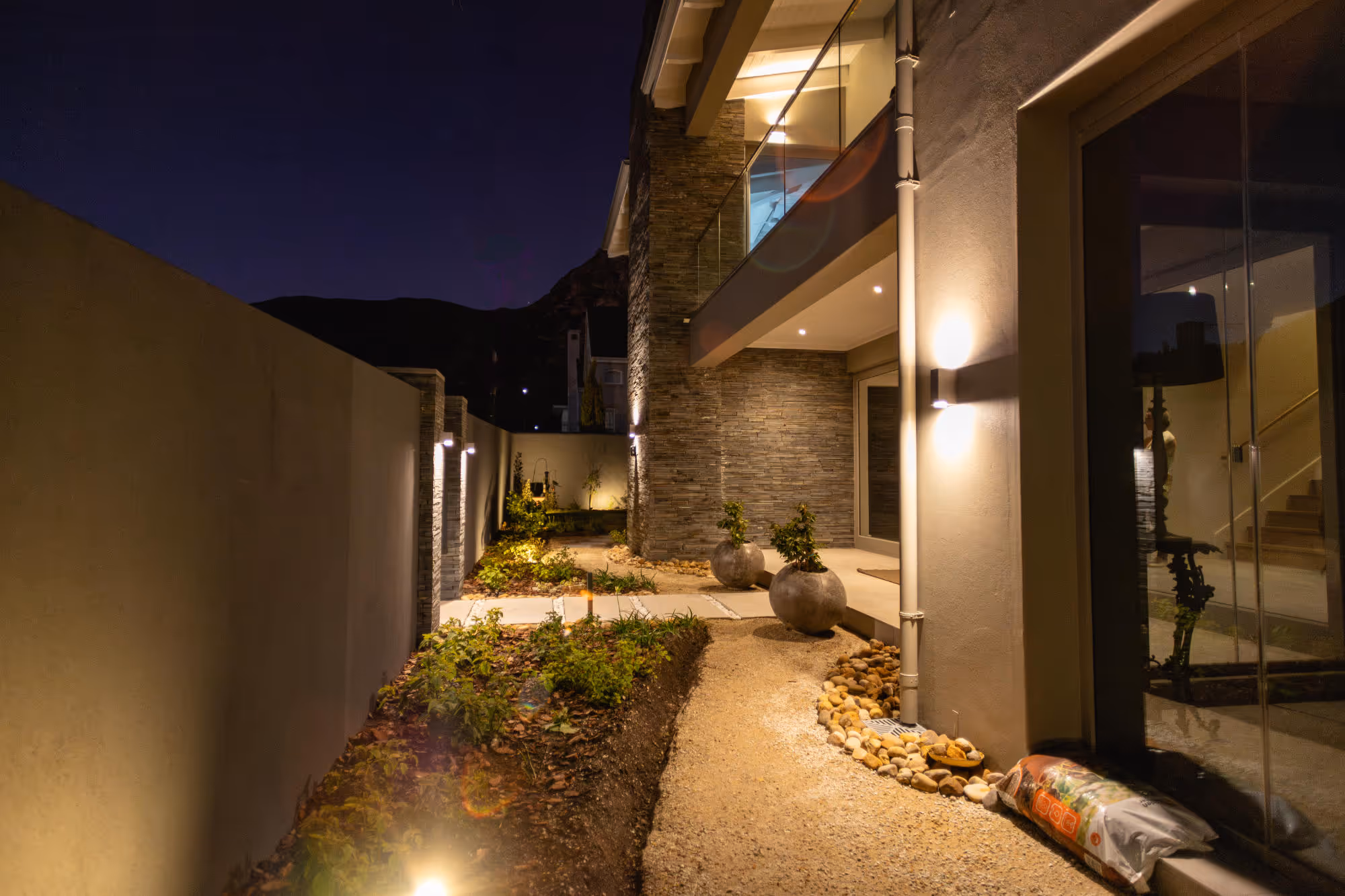 Modern house exterior at night with illuminated stone walls, potted plants, gravel pathway, and garden beds along a concrete fence. Work by Wricon construction.