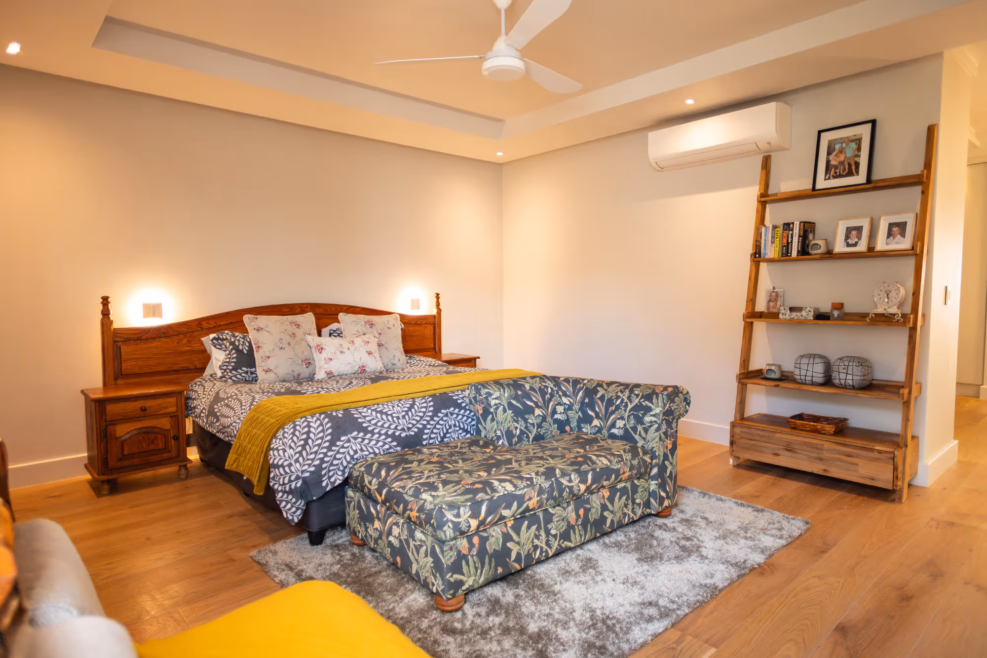 Bedroom with a wooden bed featuring patterned bedding and floral cushions, a matching floral loveseat on a gray rug, wooden nightstands, and a wooden ladder bookshelf with framed photos and books. Work by Wricon construction.