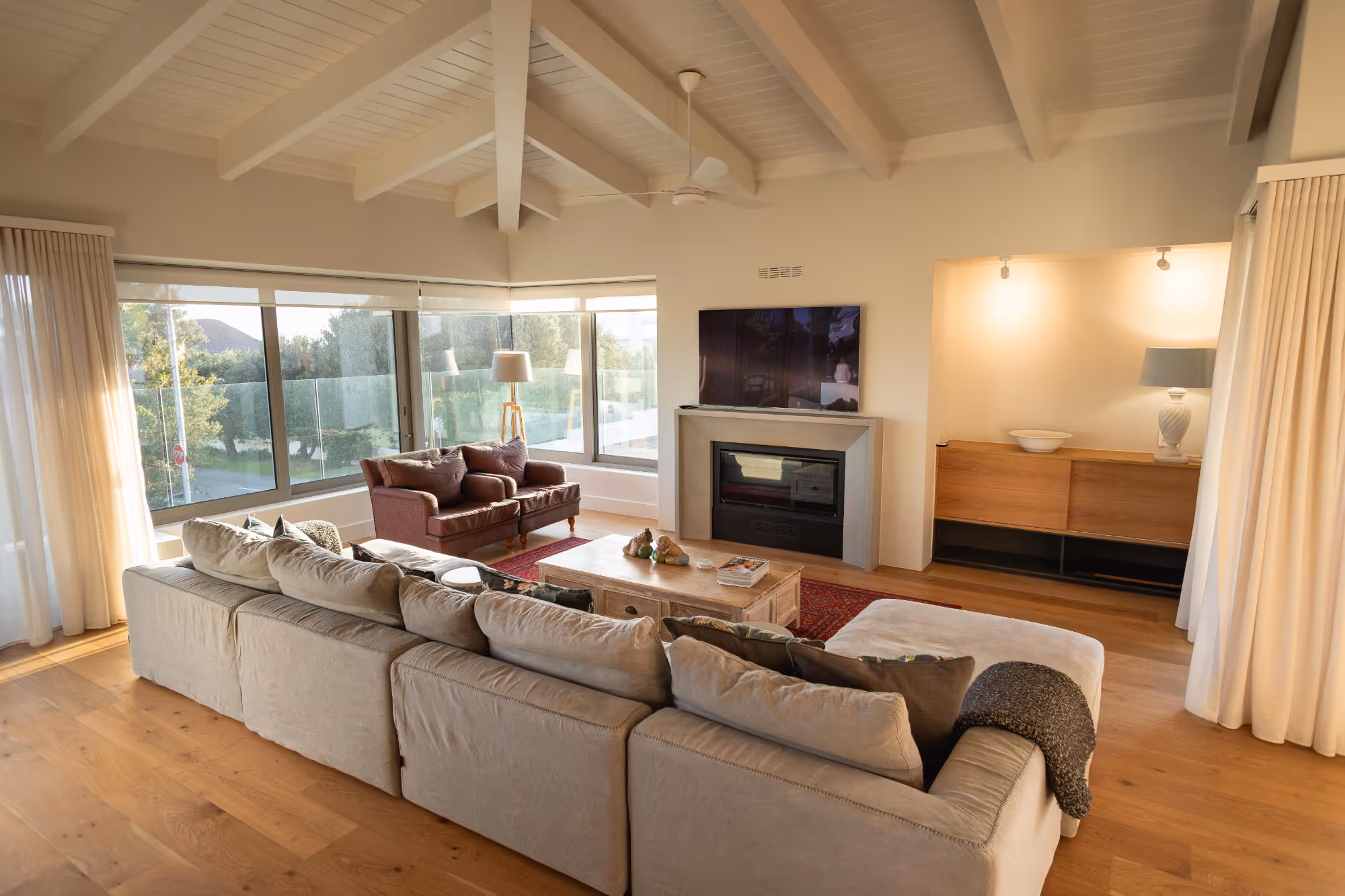 Living room with beige sectional sofa, two brown armchairs, wooden coffee table, fireplace with TV above, and large windows with natural light. Work by Wricon construction.