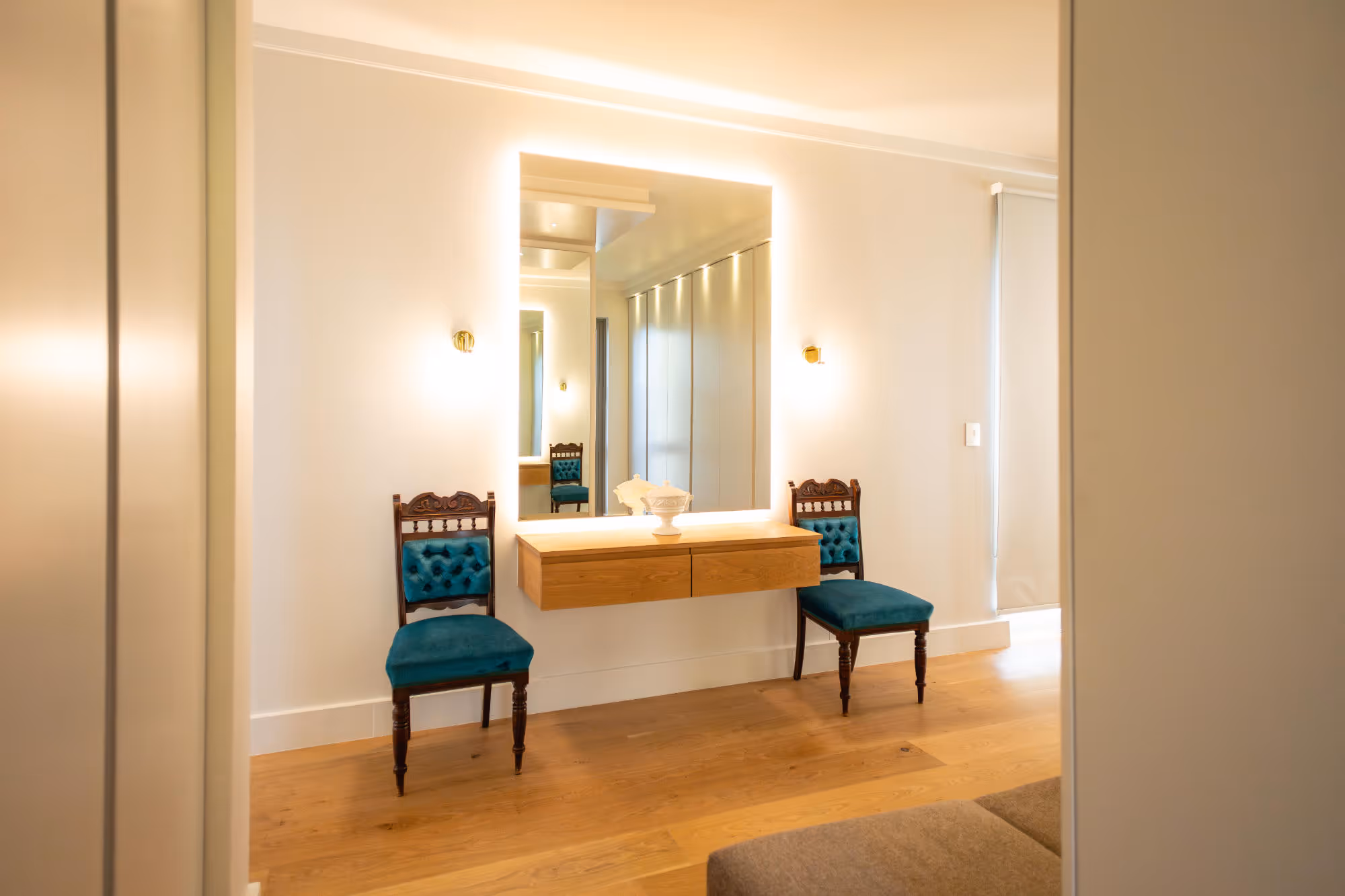 Modern vanity area with glowing rectangular mirror, floating wooden drawers, and two antique wooden chairs with teal cushions. Work by Wricon construction.