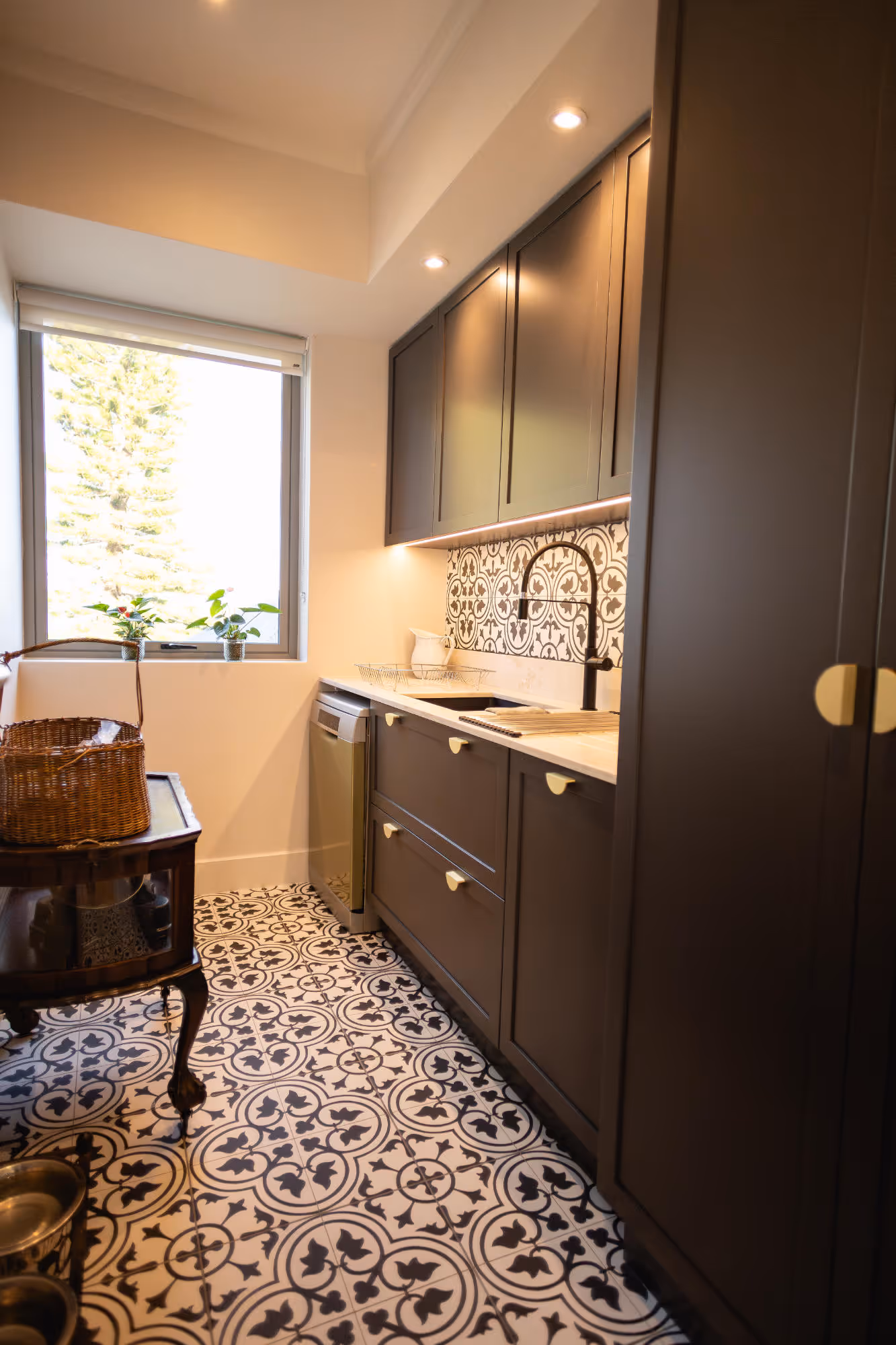 Narrow kitchen with dark cabinets, patterned black-and-white floor tiles, a window with potted plants, and under-cabinet lighting. Work by Wricon construction.