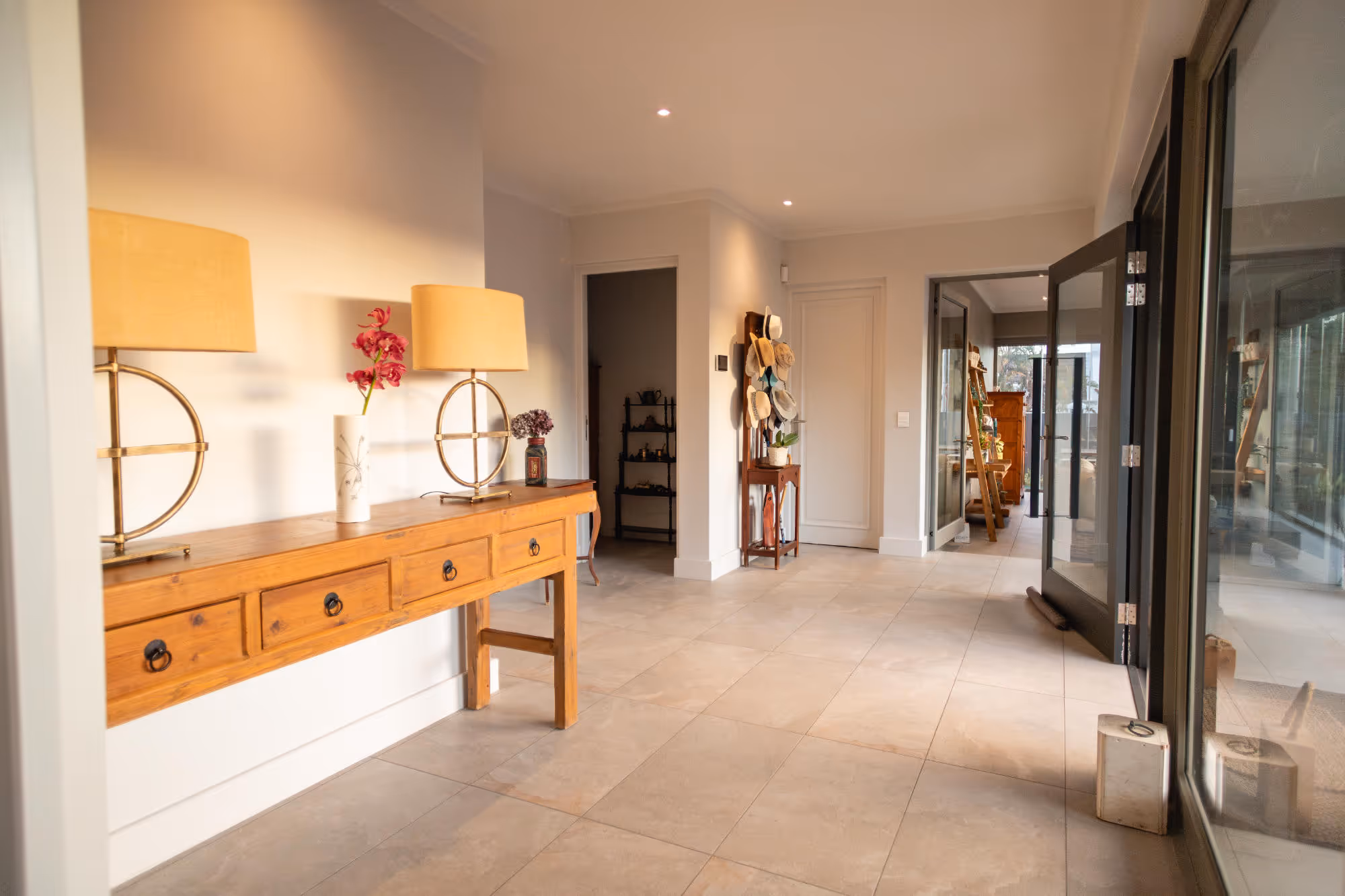 Entry hallway with wooden console table holding two round gold lamps and flower vases, hat rack, and open glass door to a bright interior space.  Work by Wricon construction.