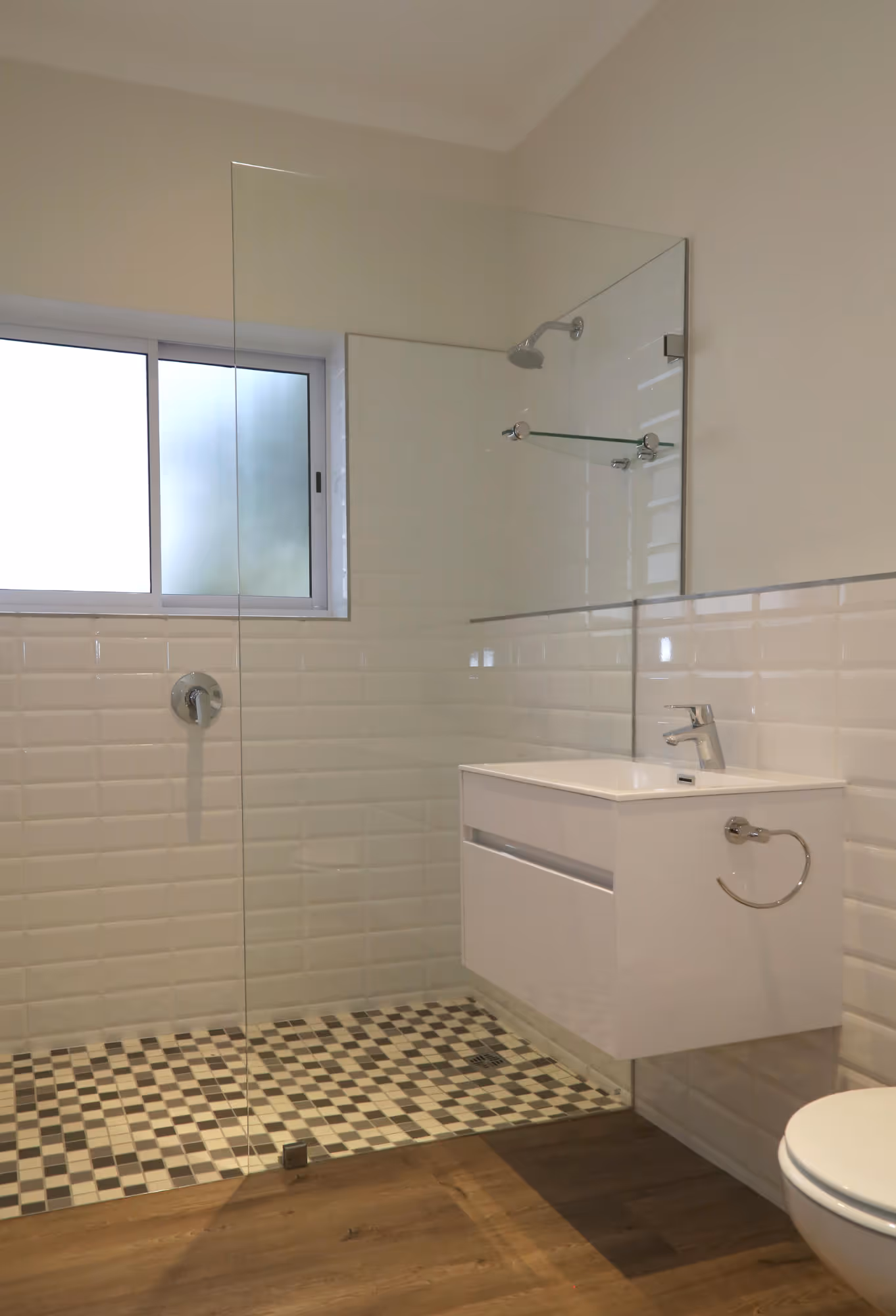 Modern bathroom with walk-in shower featuring glass partition, checkered floor tiles, white wall tiles, floating sink, and wooden floor. Work by Wricon construction.