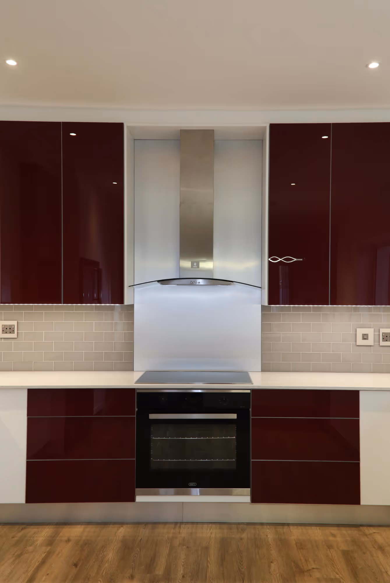 Modern kitchen with a stainless steel oven and hood, burgundy glossy cabinets, white countertop, beige subway tile backsplash, and wood flooring. Work by Wricon construction.
