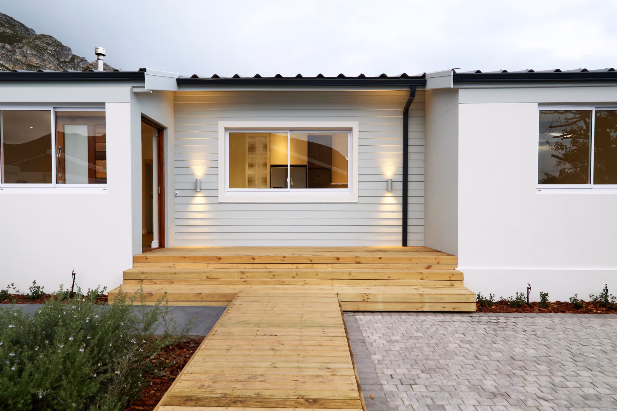 Modern single-story house front with wooden steps and pathway leading to a doorway and large windows, set against a rocky hillside. Work by Wricon construction.