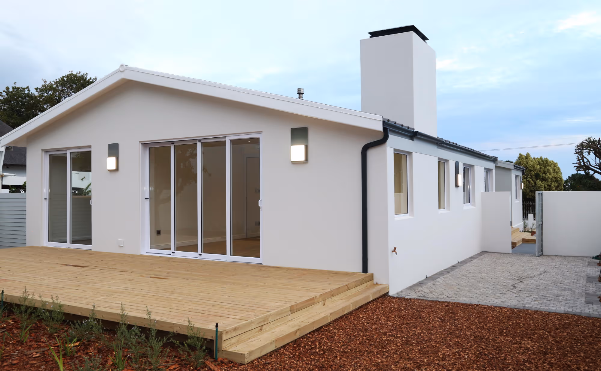 Modern white single-story house with large sliding glass doors opening to a wooden deck and a paved driveway on the side. Work by Wricon construction.