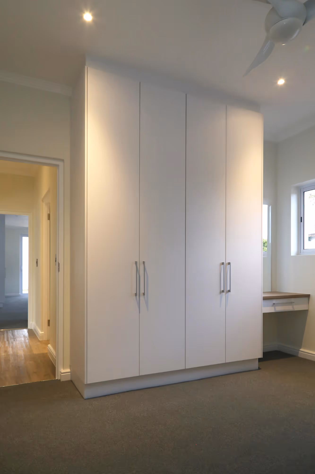 Minimalist room with a tall white wardrobe with four doors and a small built-in desk under a window. Work by Wricon construction.