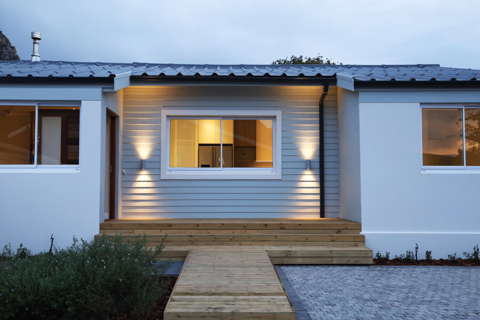 Modern house exterior at dusk with wooden steps, light gray siding, and warm interior lights visible through windows. Work by Wricon construction.