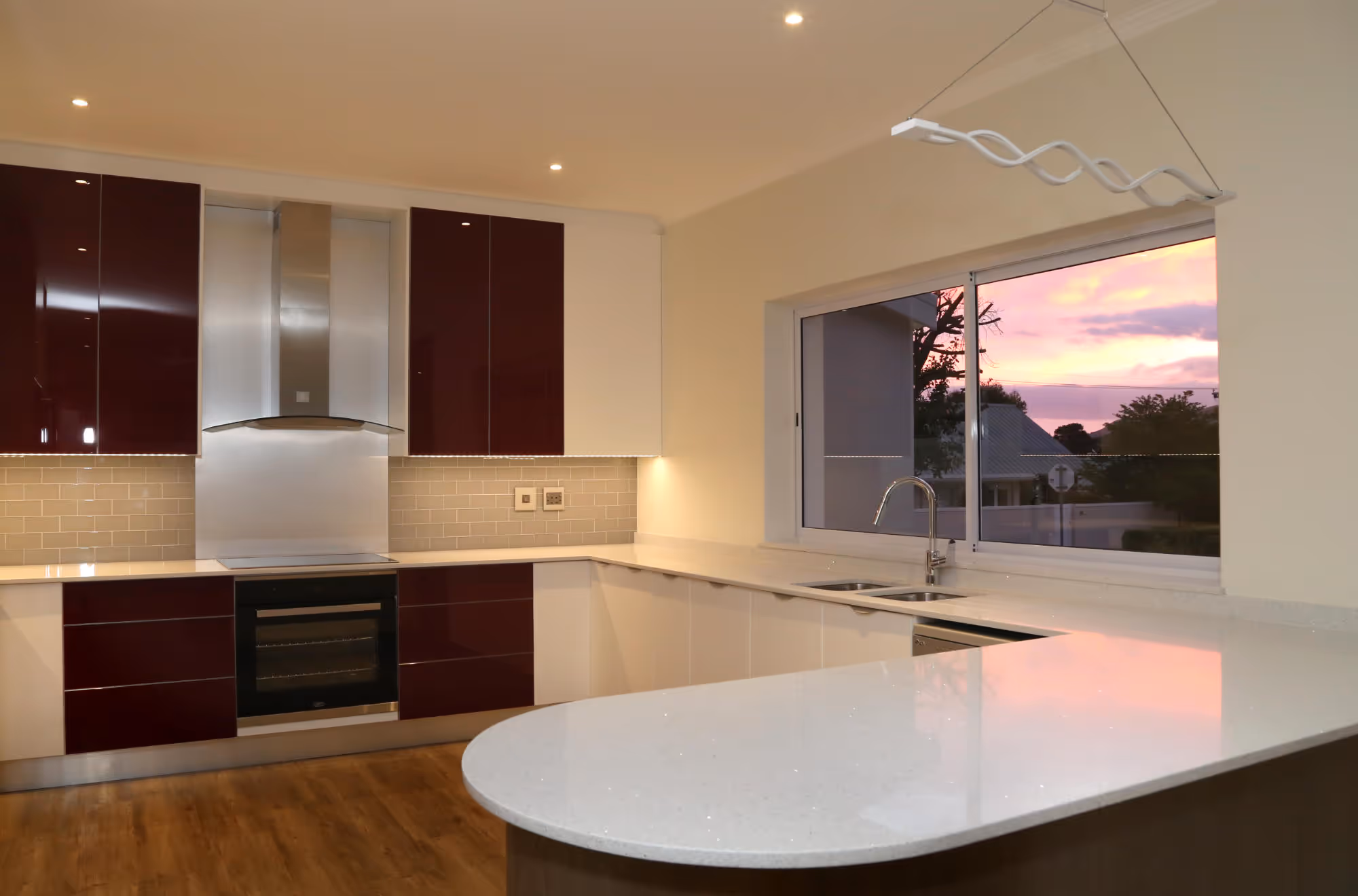 Modern kitchen with glossy maroon and white cabinets, white countertops, stainless steel oven and hood, large window showing a colorful sunset. Work by Wricon construction.