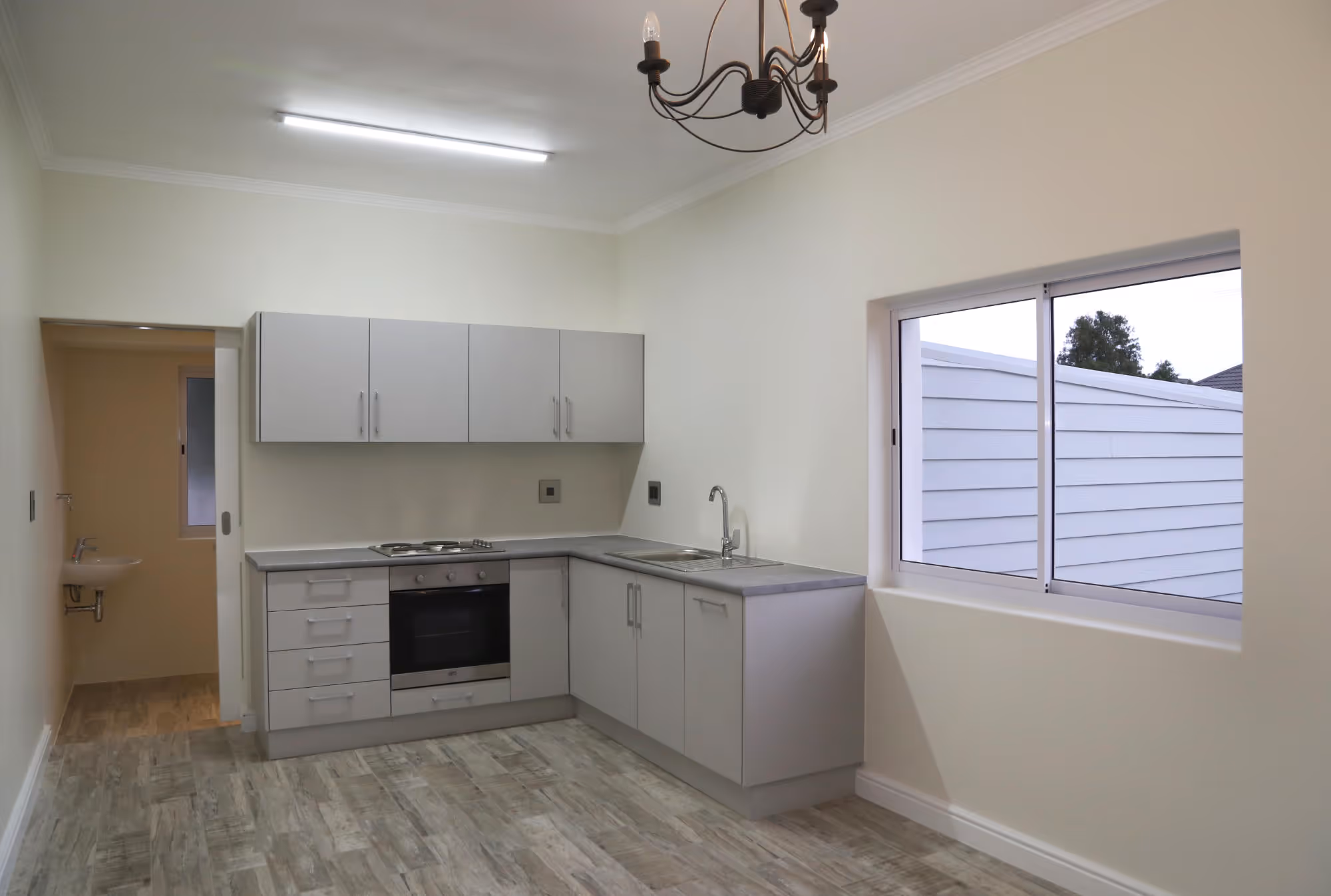 Small clean kitchen with gray cabinets, built-in oven, stovetop, sink, and a window overlooking a white fence. Work by Wricon construction.