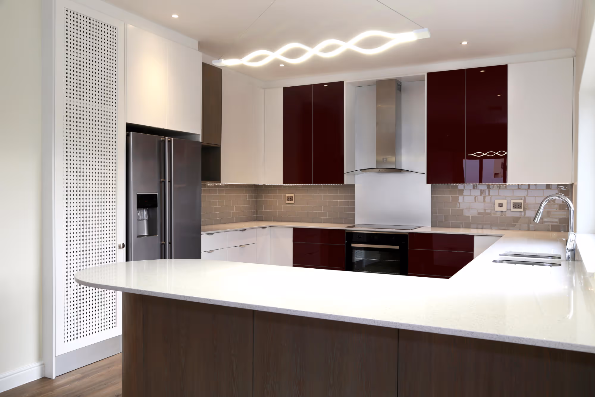 Modern kitchen with white and dark red cabinets, a large white countertop island, stainless steel refrigerator, stovetop oven, and a unique wave-shaped ceiling light. Work by Wricon construction.