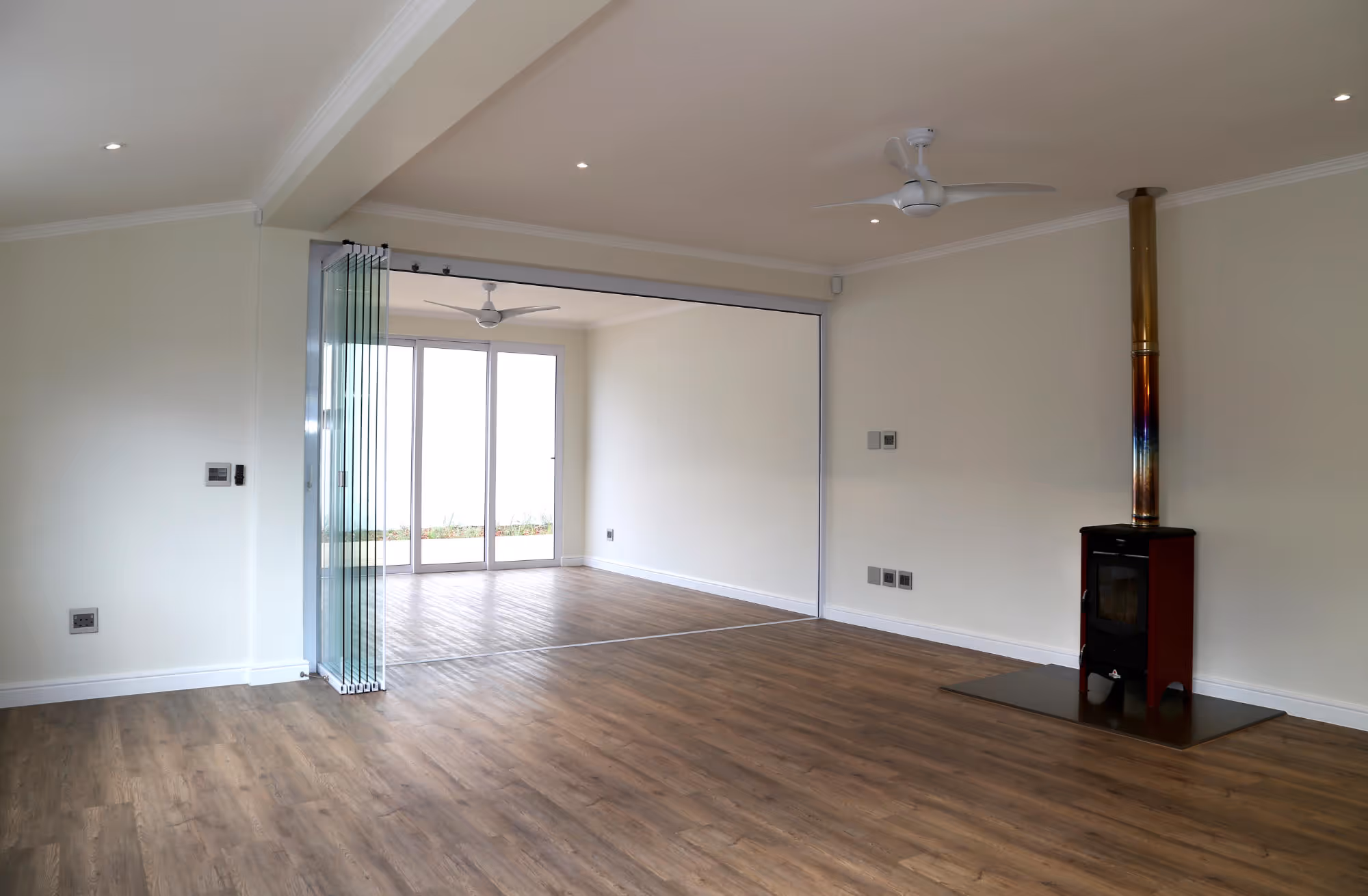 Empty modern living room with wood flooring, glass folding doors leading to an adjoining room, ceiling fans, and a black wood stove with a tall metallic chimney. Work by Wricon construction.